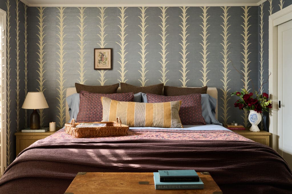 Blue bedroom with cream vertical leaf-pattern wallpaper; bed layered with striped and patterned pillows, wicker basket on the footboard, and a vase of red flowers on the right bedside table.