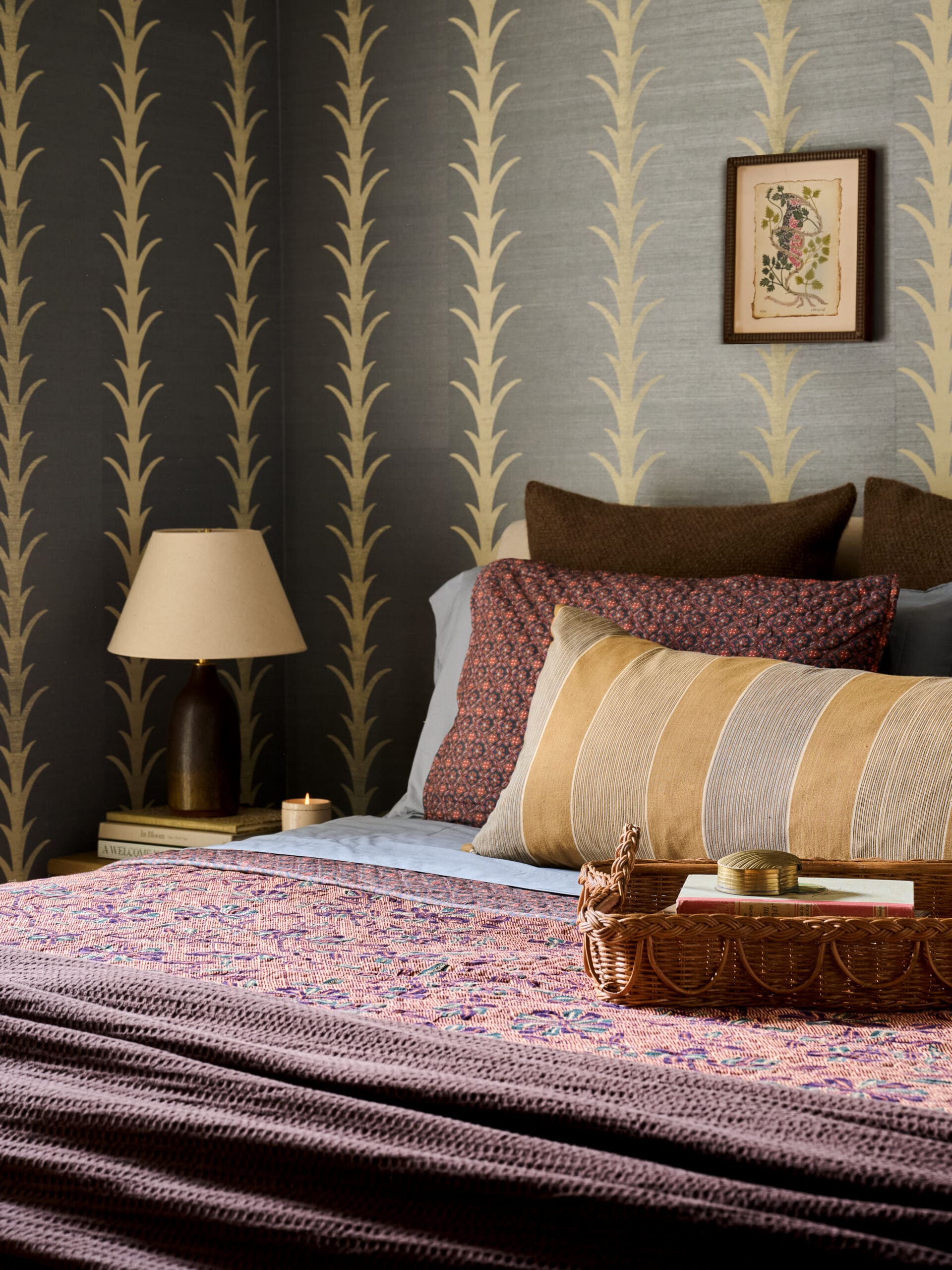 Bedroom corner with dark gold-patterned wallpaper, a framed botanical print, and a bed piled with striped and floral pillows near a nightstand with a lamp.
