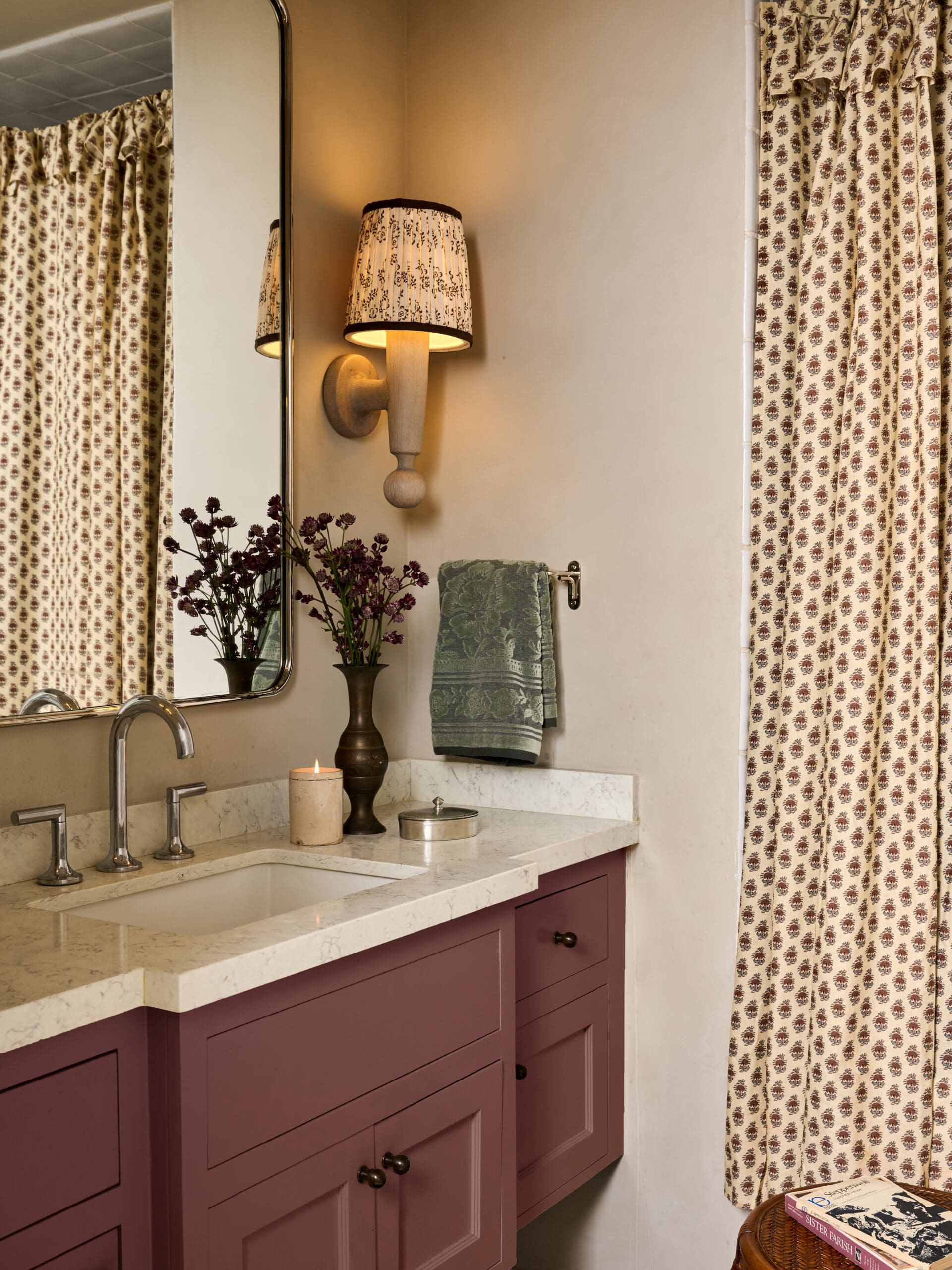 Pink bathroom vanity with a marble countertop, dual chrome faucets, and lower drawers. A wall lamp with a patterned shade is above, reflected in the mirror.