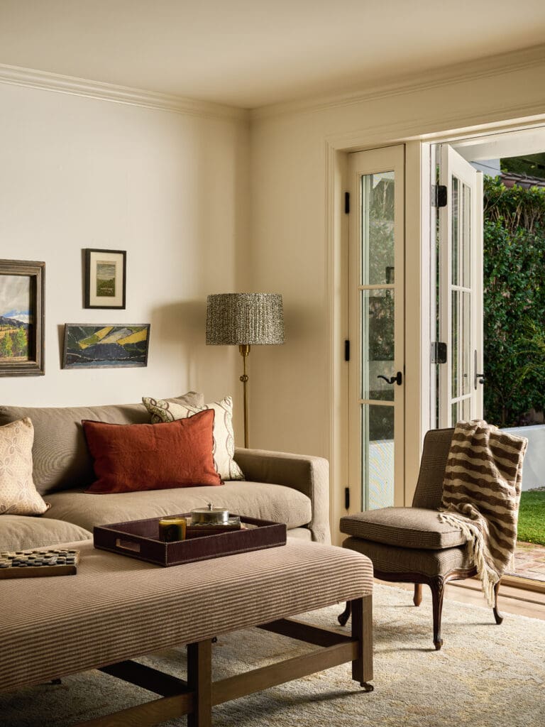 Cozy living room with a beige striped sofa, red and patterned pillows, wall art, and open French doors to a lush garden outside.