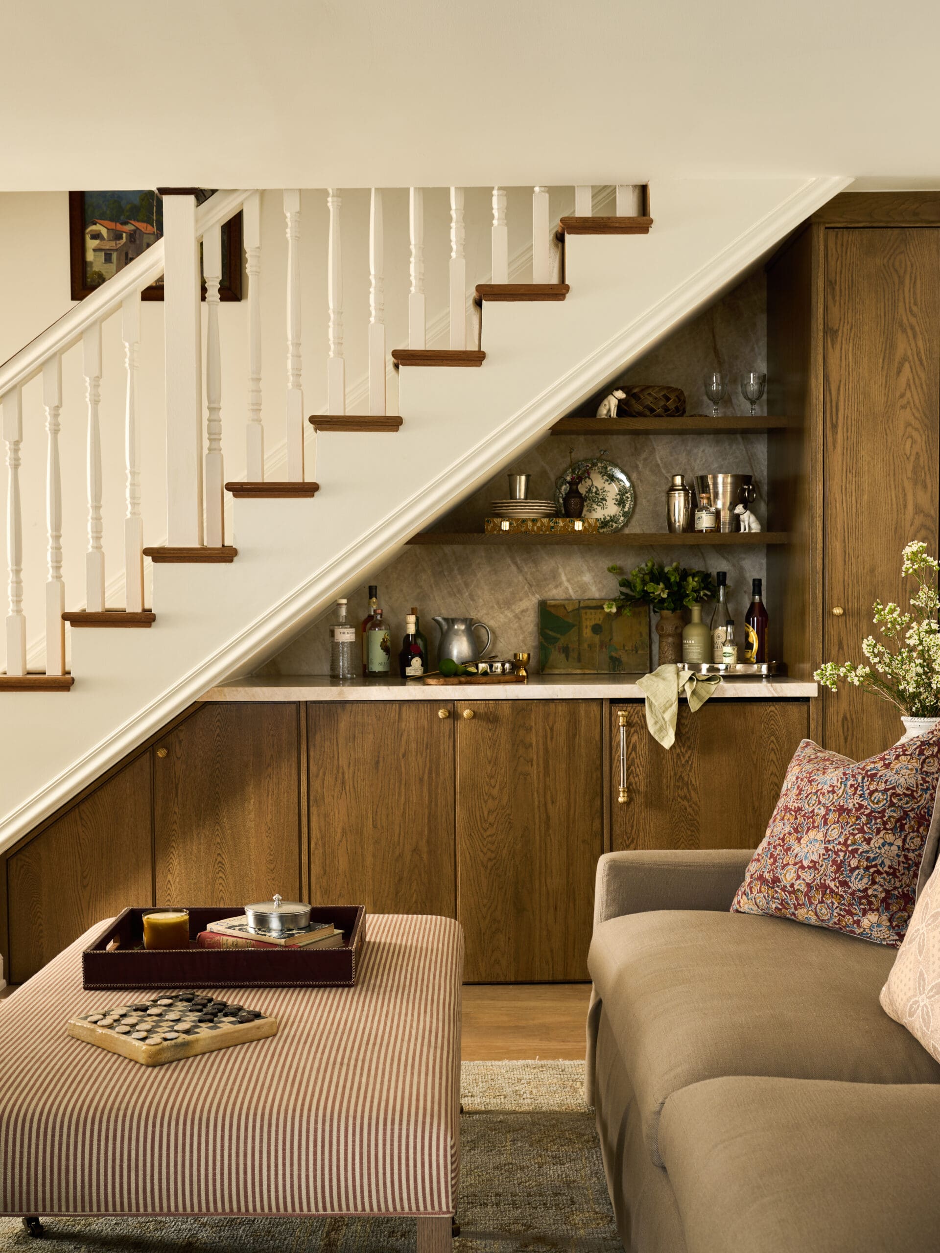 Cozy living area under a staircase with white railings, open shelves holding glassware and bottles, and a beige sofa nearby.