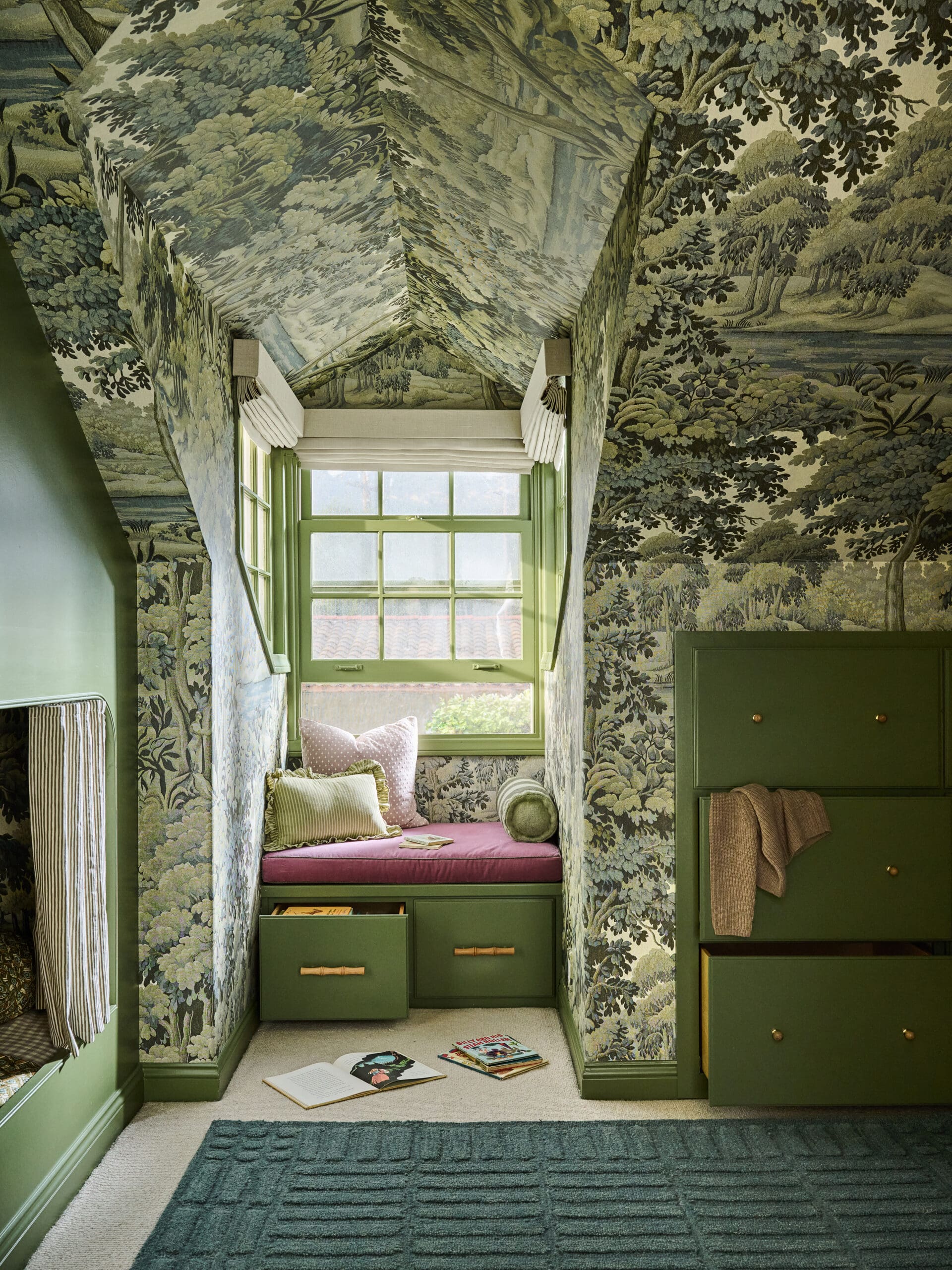Cozy window seat with pink cushion and green built‑in drawers, surrounded by blue-green toile wallpaper and a large window.
