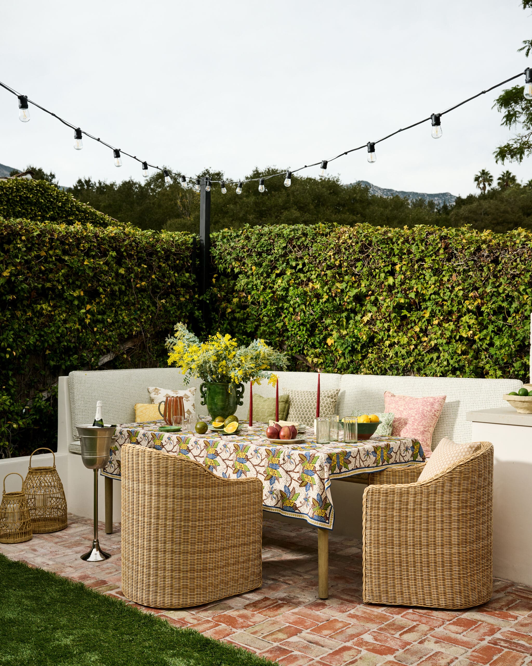 Outdoor dining setup with a patterned tablecloth, wicker chairs, and string lights above against a green hedge backdrop.