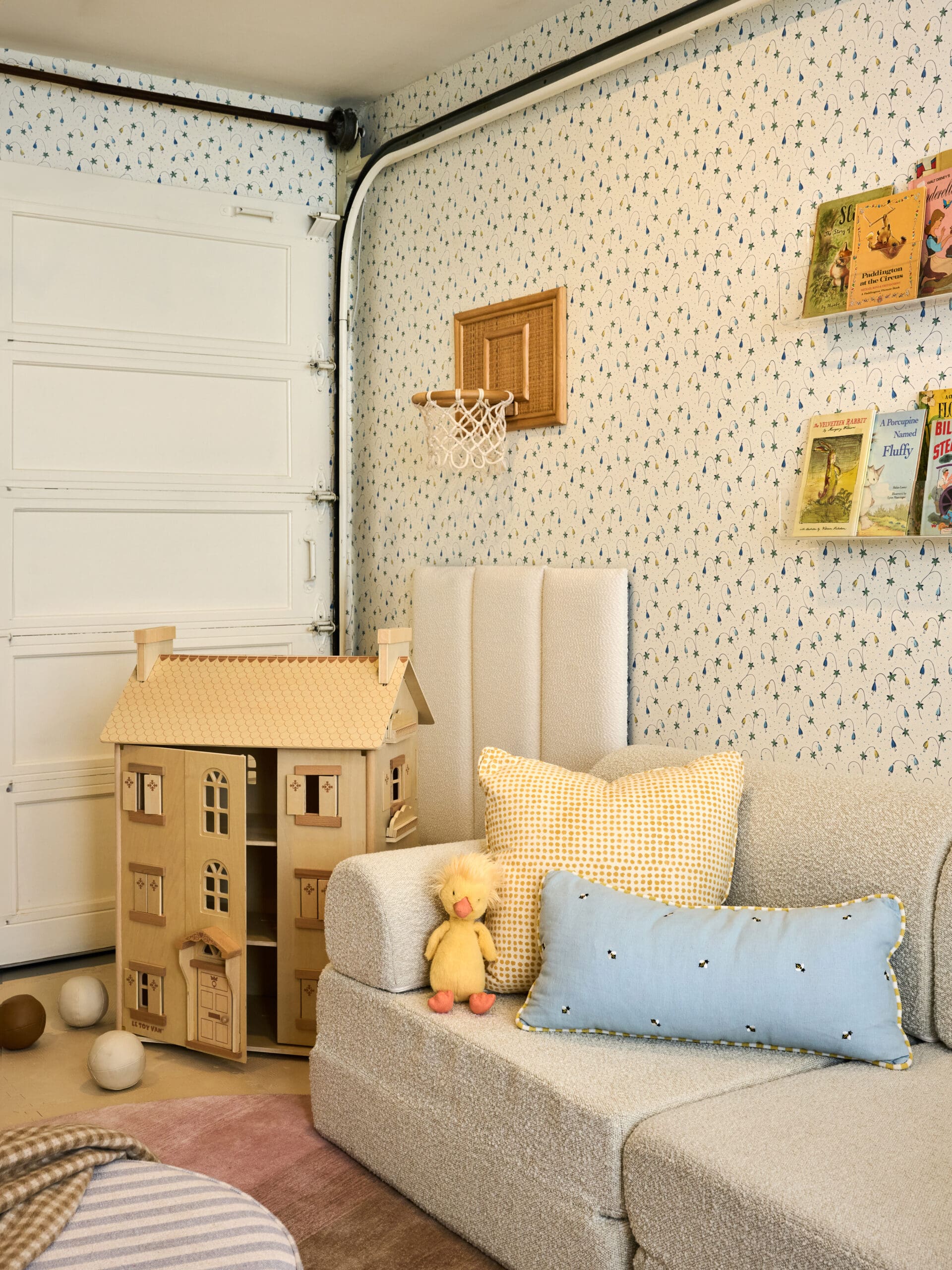 Playroom corner with a wooden dollhouse, a gray sofa, yellow patterned cushions, and a wall shelf of children’s books against patterned wallpaper.