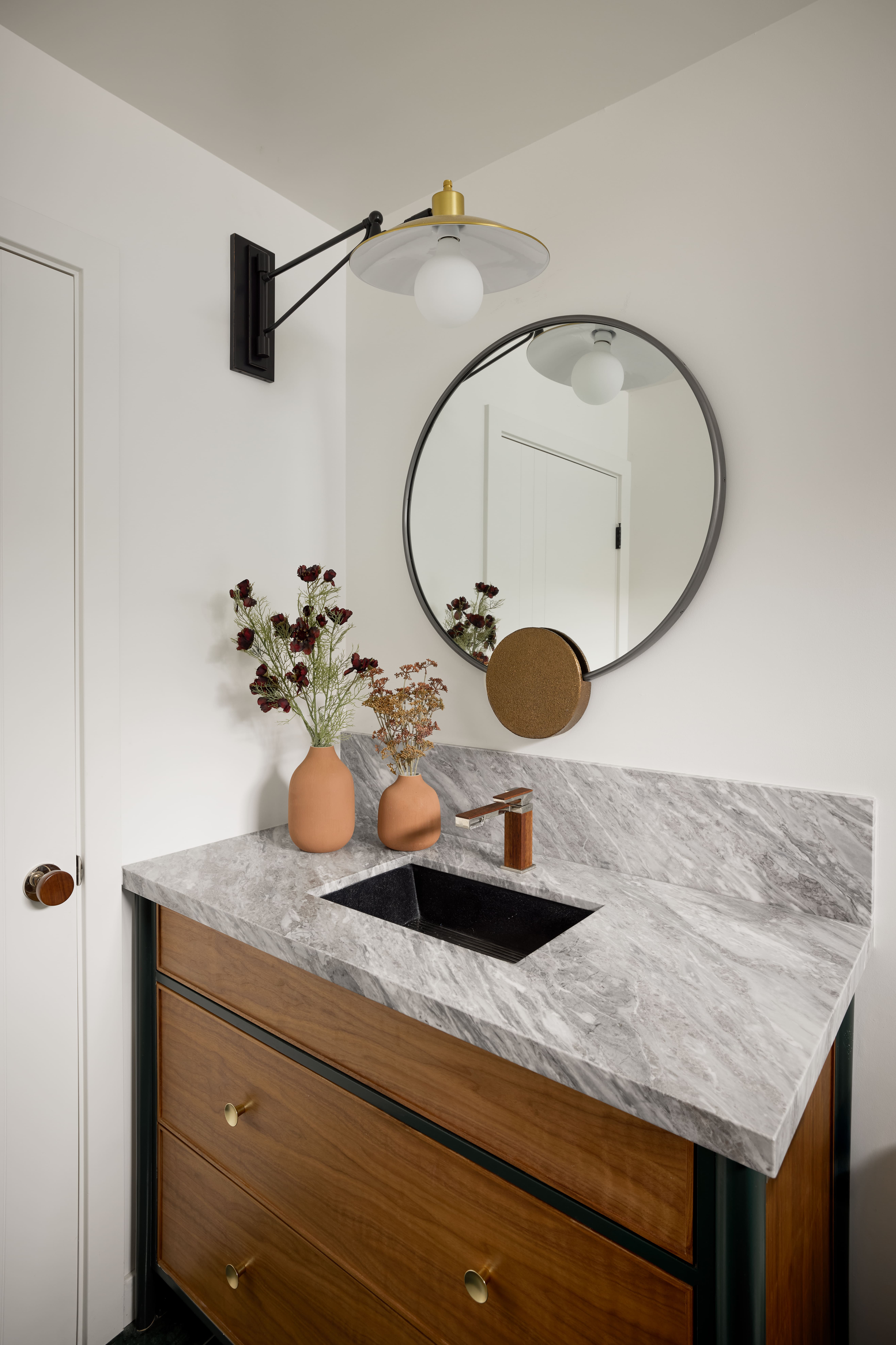 Bathroom vanity with gray marble countertop, wood drawers, round mirror, and modern wall-mounted light fixture with gold accent.