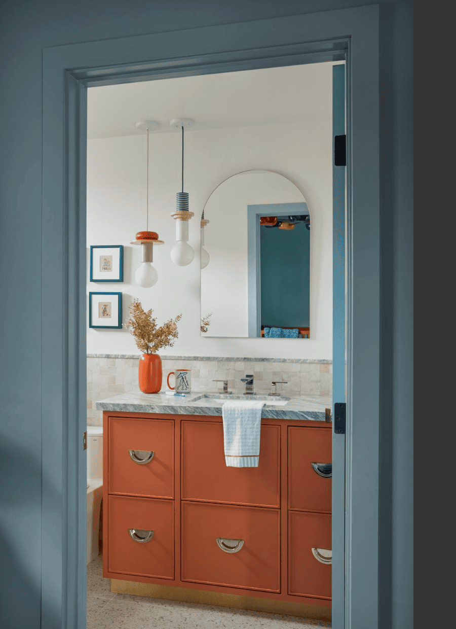 Bright bathroom with an orange six-drawer vanity, marble countertop, towel hanging over the sink, a vase of dried flowers, and a round mirror.