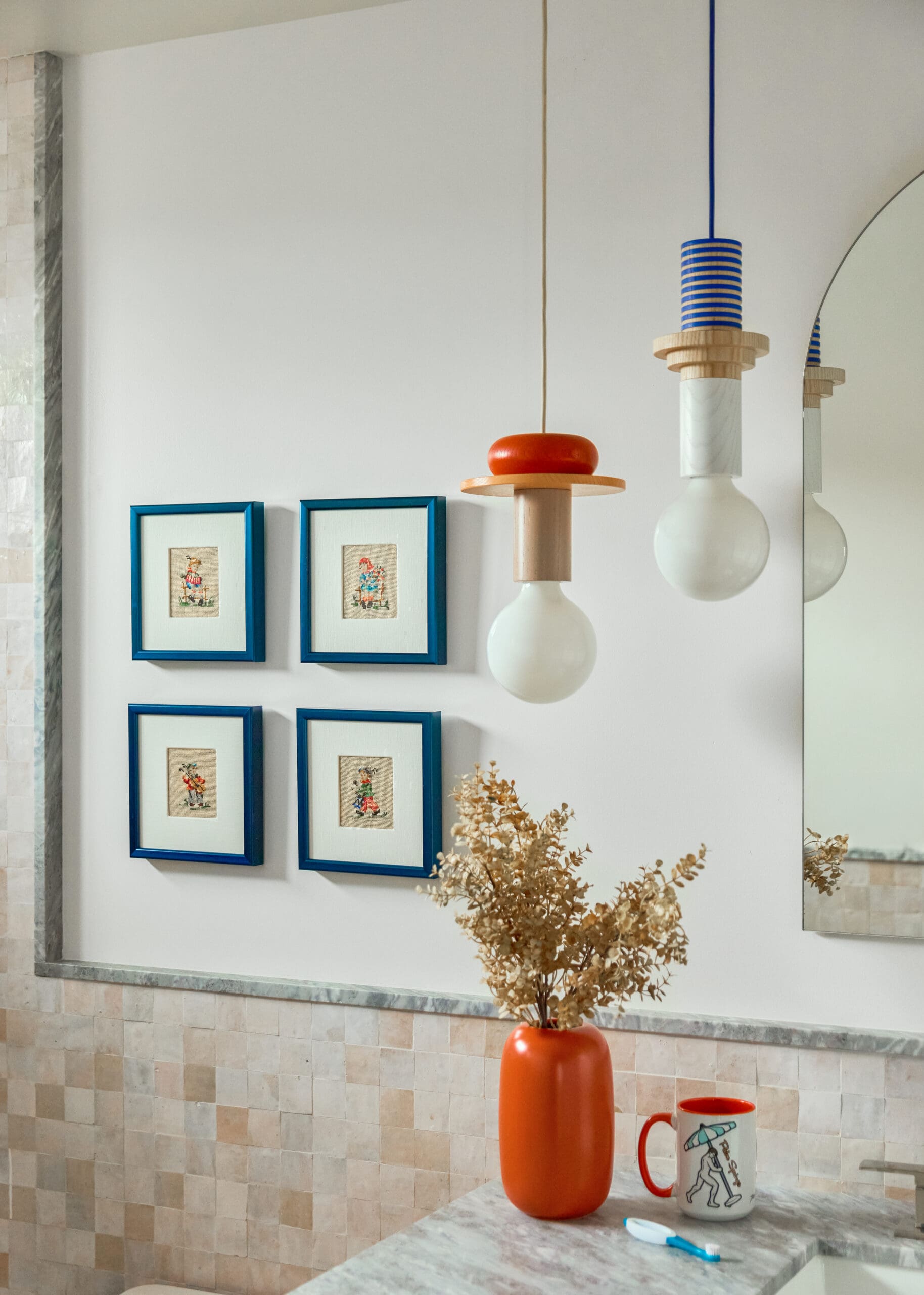Blue-framed small prints on a white wall with an orange vase holding dried branches, plus a countertop toothbrush and a mug with a figure drawing.