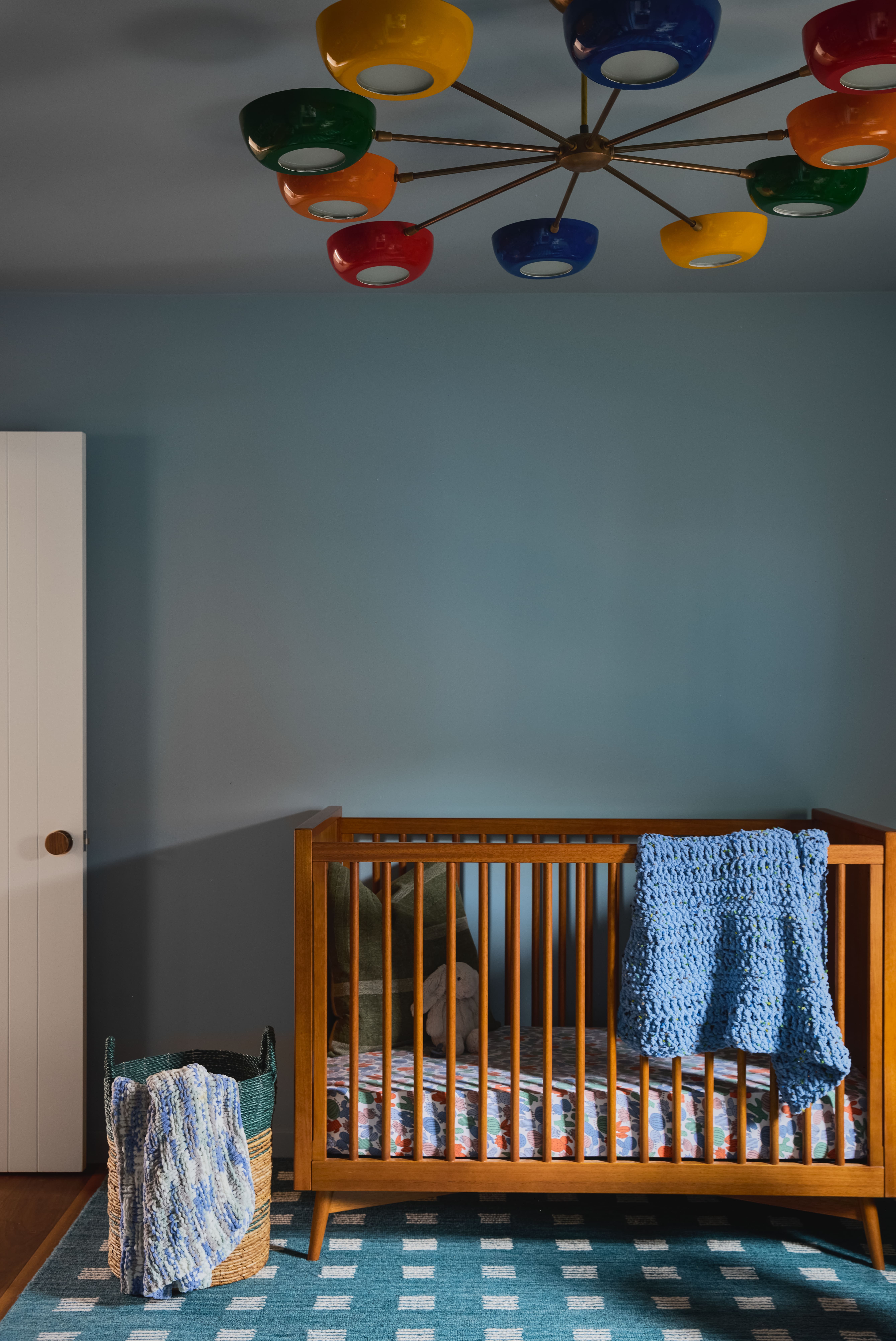 Wooden baby crib against a blue wall, with a blue-and-white patterned rug, a wicker basket with a blue blanket, and a colorful globe chandelier overhead.