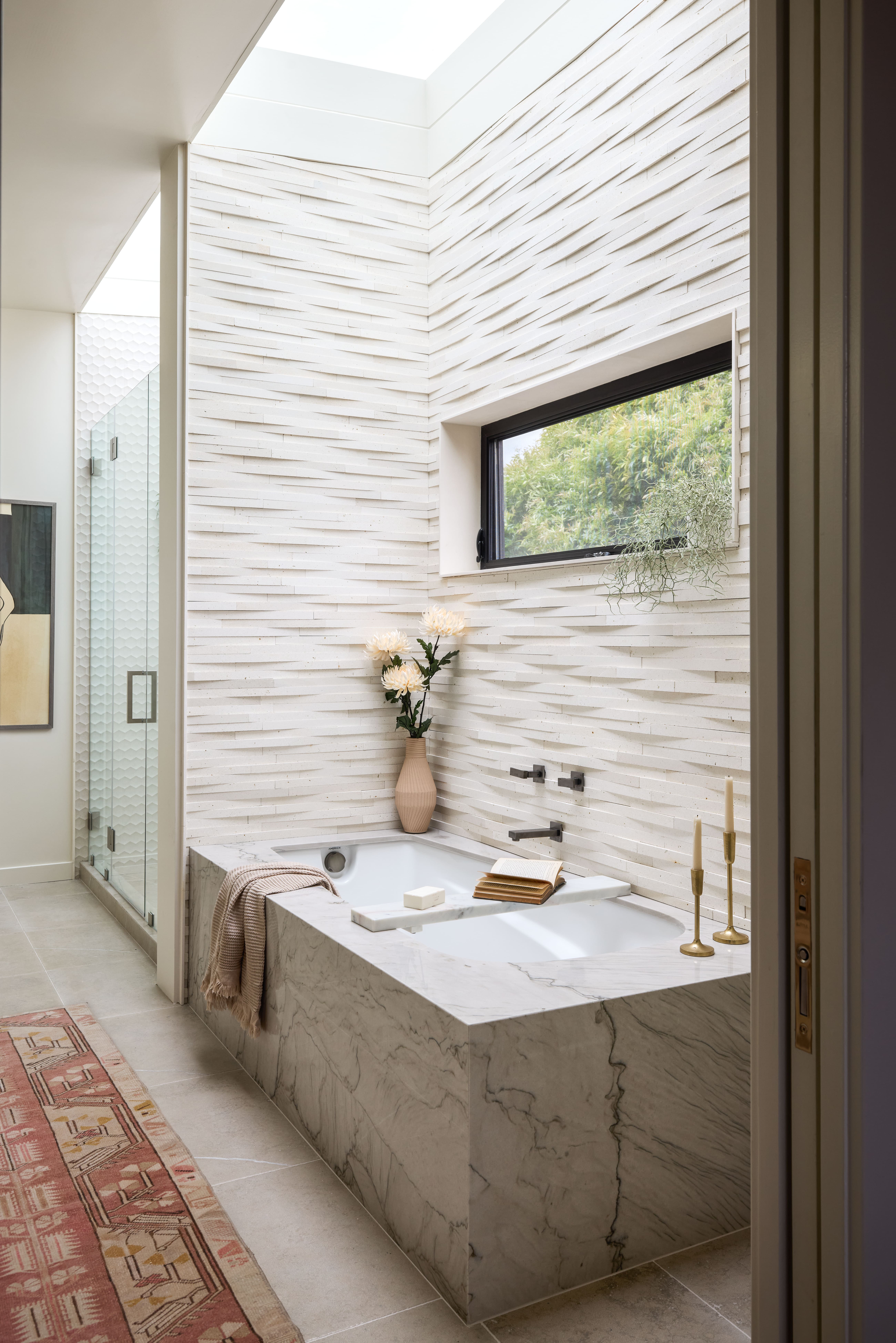 Marble bathtub built into a marble surround with a beige towel draped over the edge in a bright bathroom with a long horizontal window and a vase of flowers nearby.