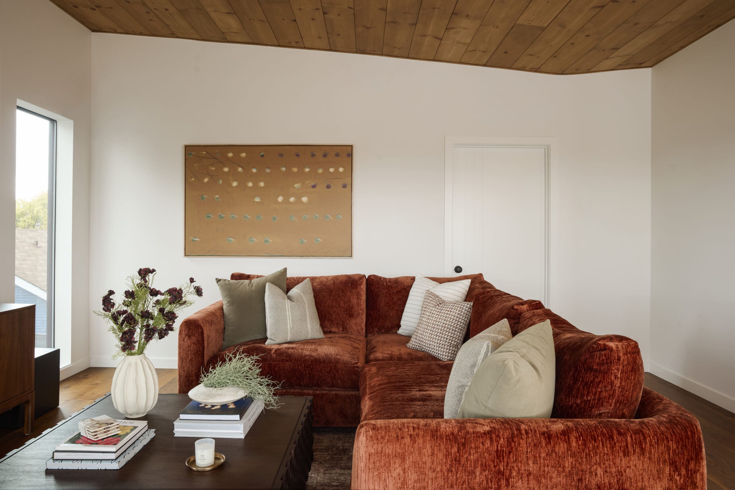 Rust velvet sofa with beige and patterned cushions in a bright living room, a dark coffee table with magazines and a white vase of dried flowers in front; framed abstract art on the wall, left window, and a wooden ceiling above.