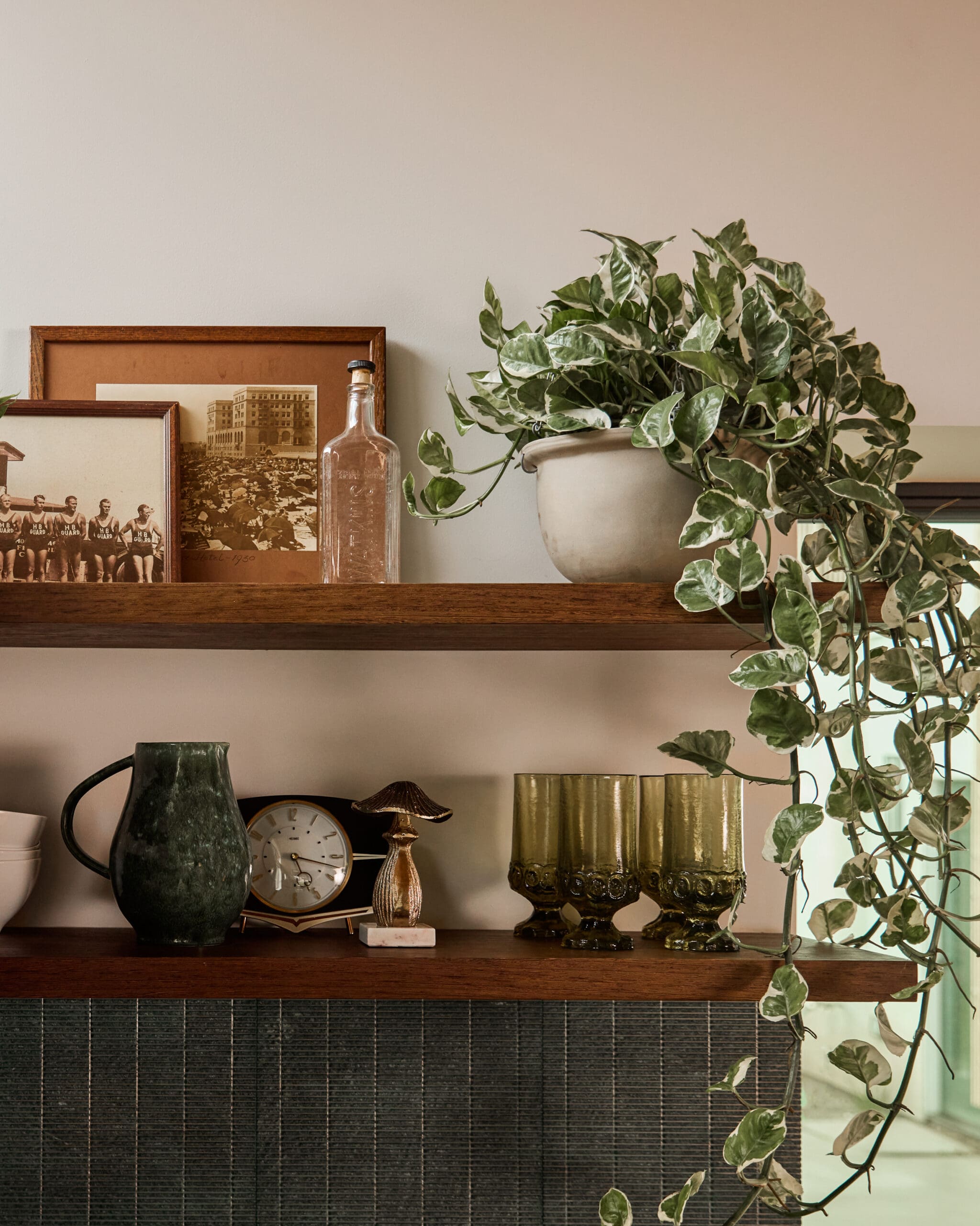 Shelf with vintage framed photos, a glass bottle, clock, and brass accents beside a trailing ivy plant on a beige wall.
