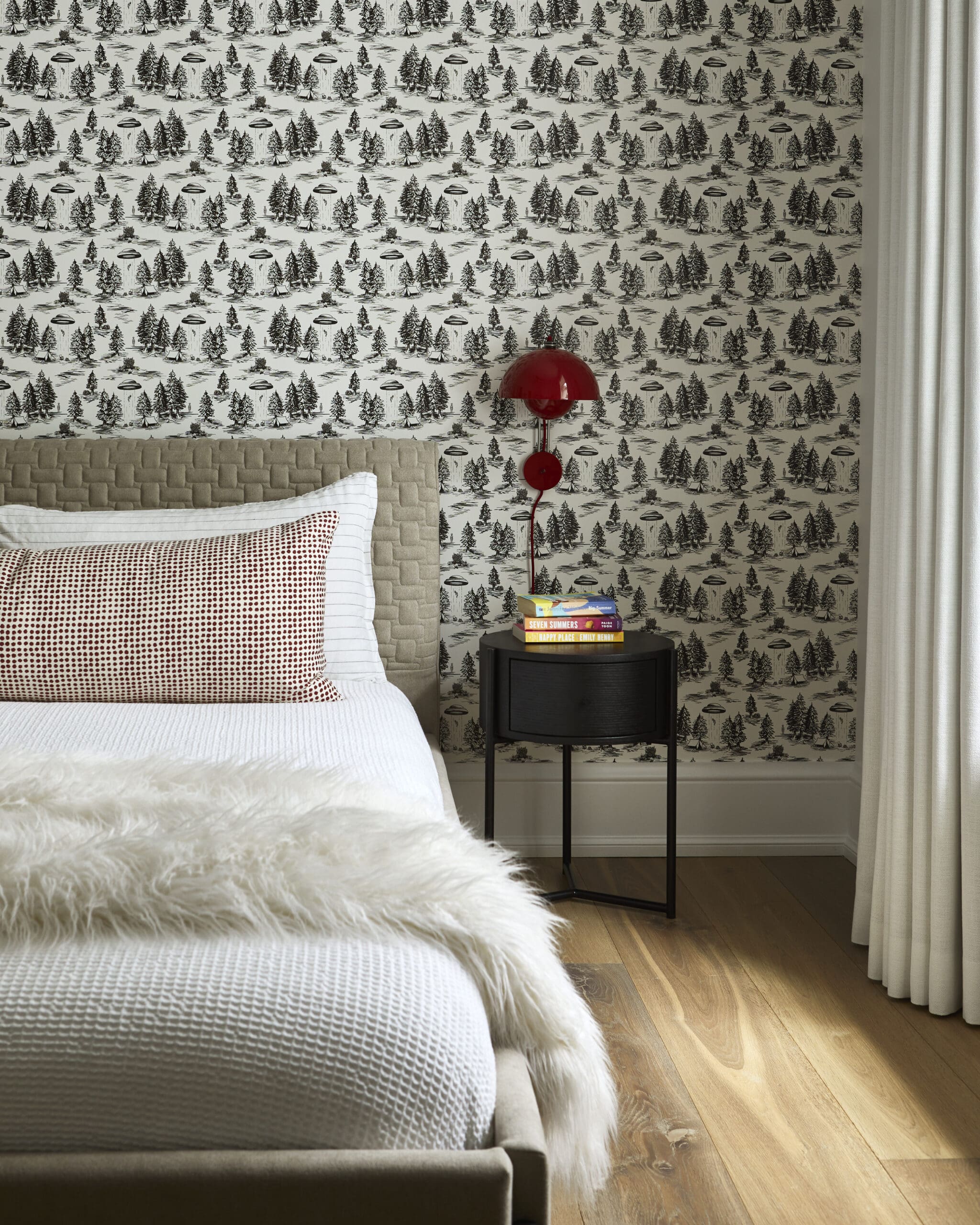 Bedroom scene with a beige upholstered bed, white bedding, and a fluffy white throw against a black-and-white forest wallpapered wall.