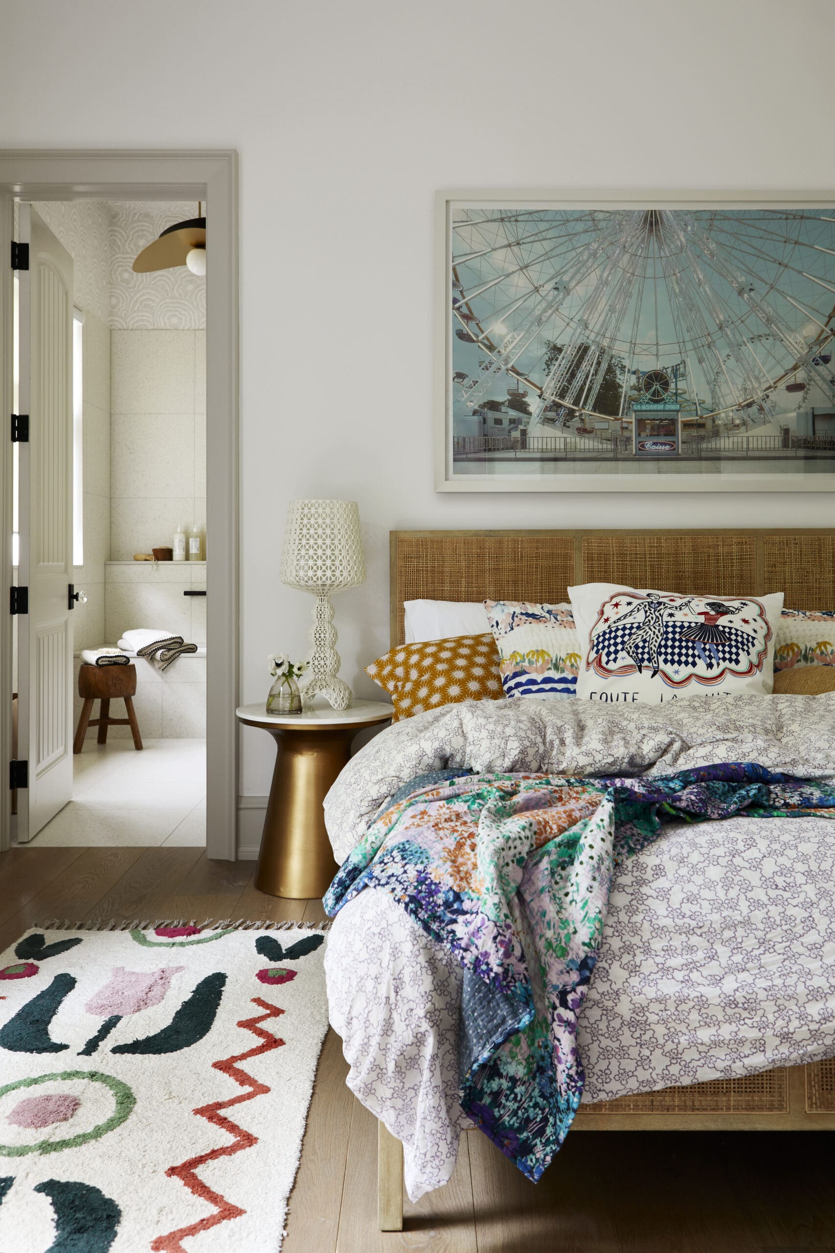 Bedroom with a wicker headboard, colorful quilts and pillows, and a large Ferris wheel photo on the wall; bathroom door open to the left.