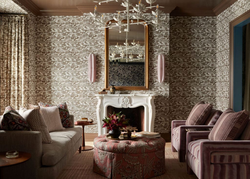 Living room with ornate patterned wallpaper, white fireplace mantel, a gold-framed mirror, and pink upholstered seating around a tufted ottoman.