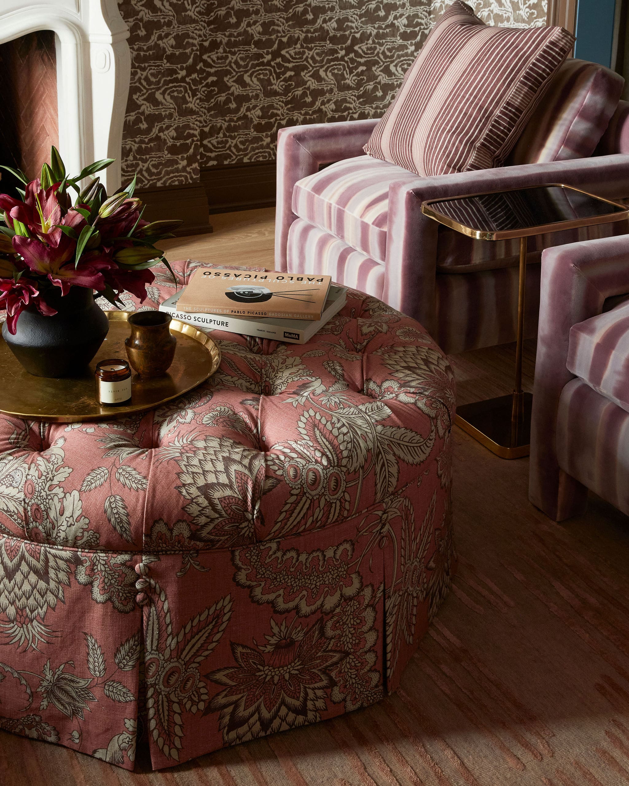 Pink patterned sofa with a striped cushion, a gold-framed side table, and a vase of red flowers on a brass tray in a luxurious living room.