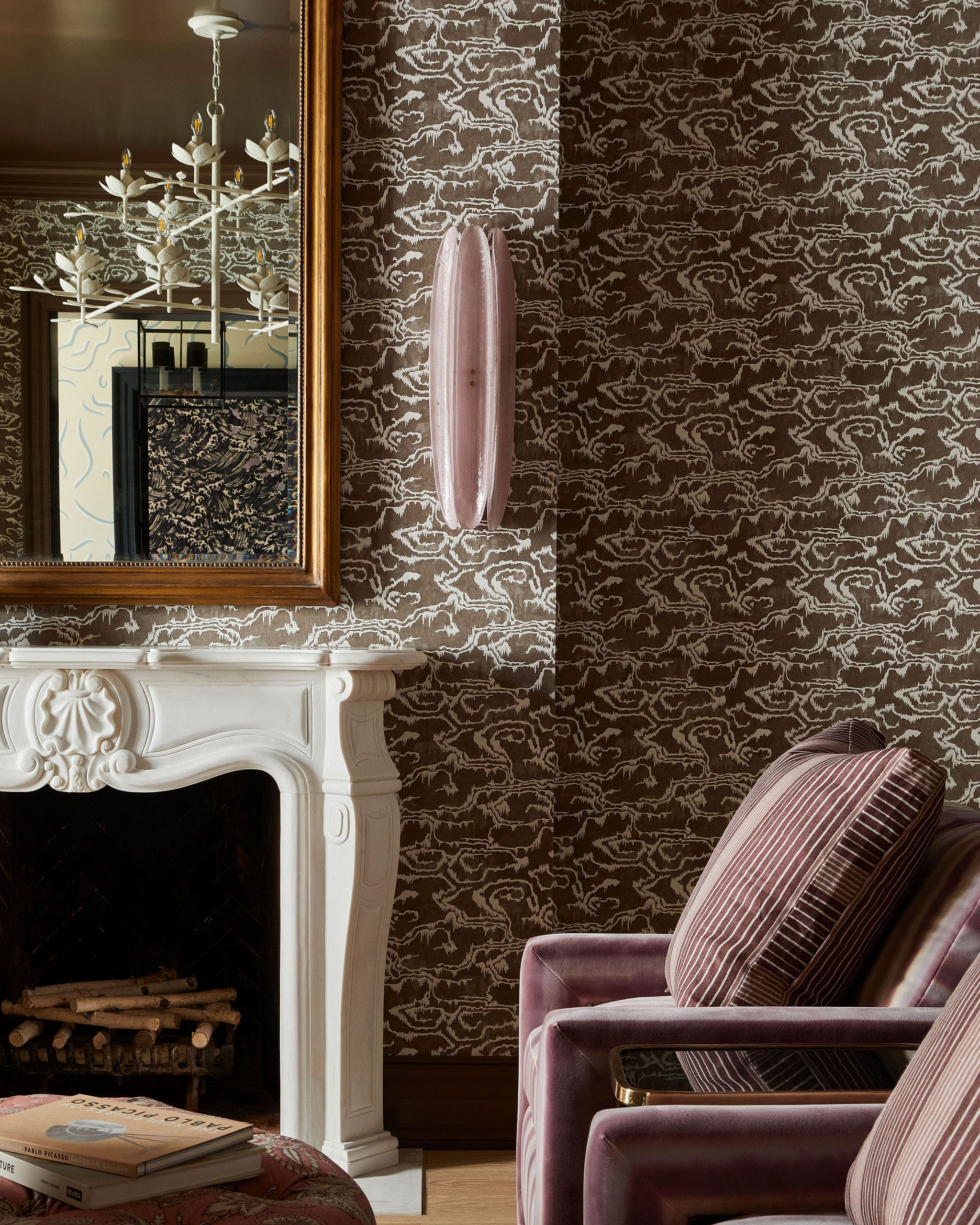 White ornate fireplace with a large wooden-framed mirror, brown patterned wallpaper, a pink velvet chair, and a stack of art books.
