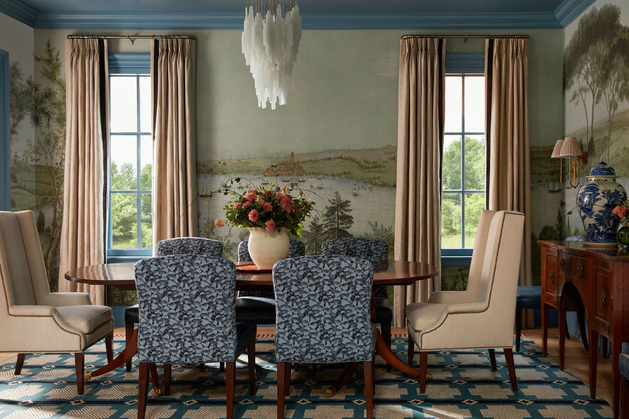 Elegant dining room with a round wooden table, blue patterned chairs, a floral centerpiece, and a white chandelier.