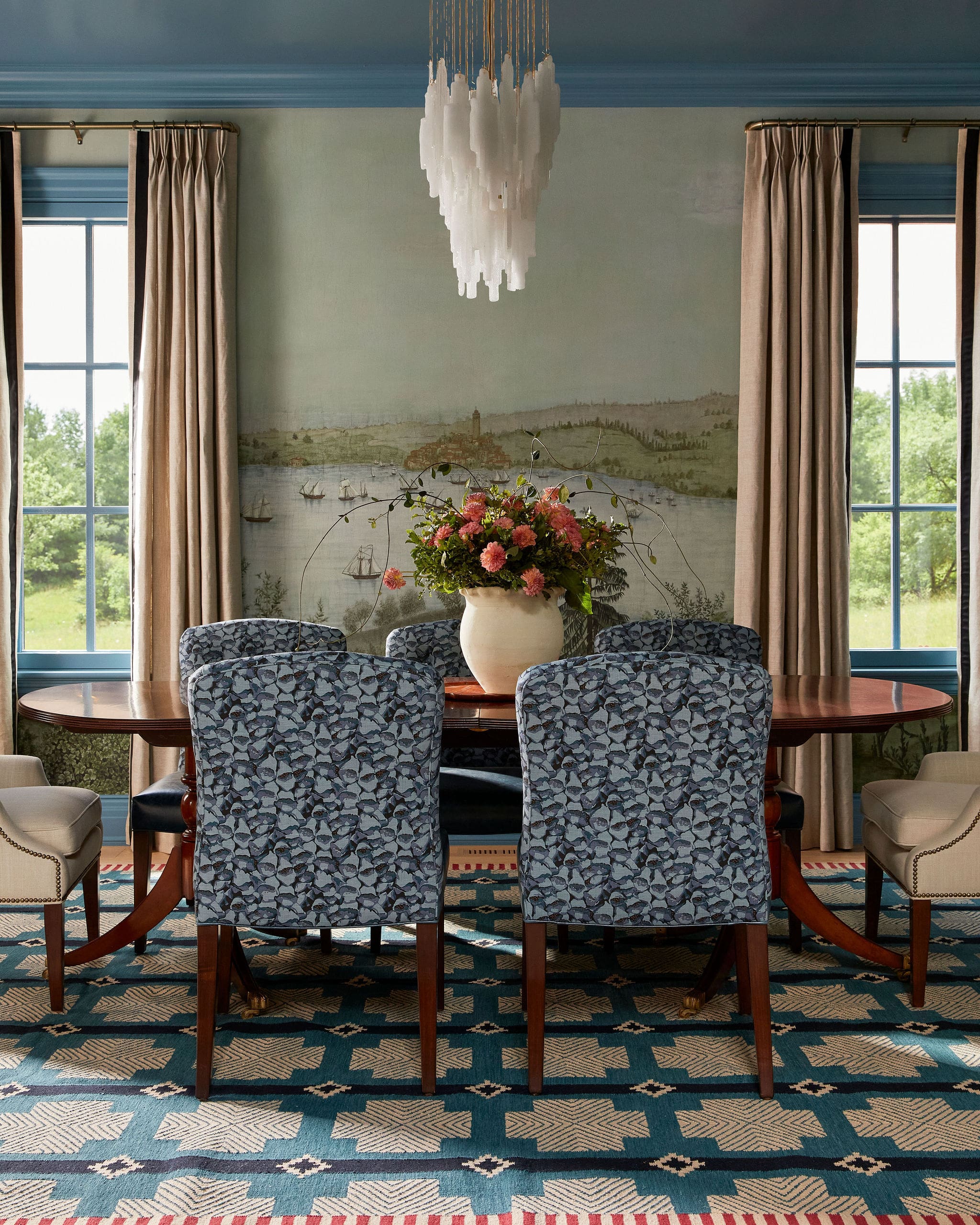 Elegant dining room scene with a round wooden table, blue patterned chairs, and a large floral centerpiece under a white chandelier.