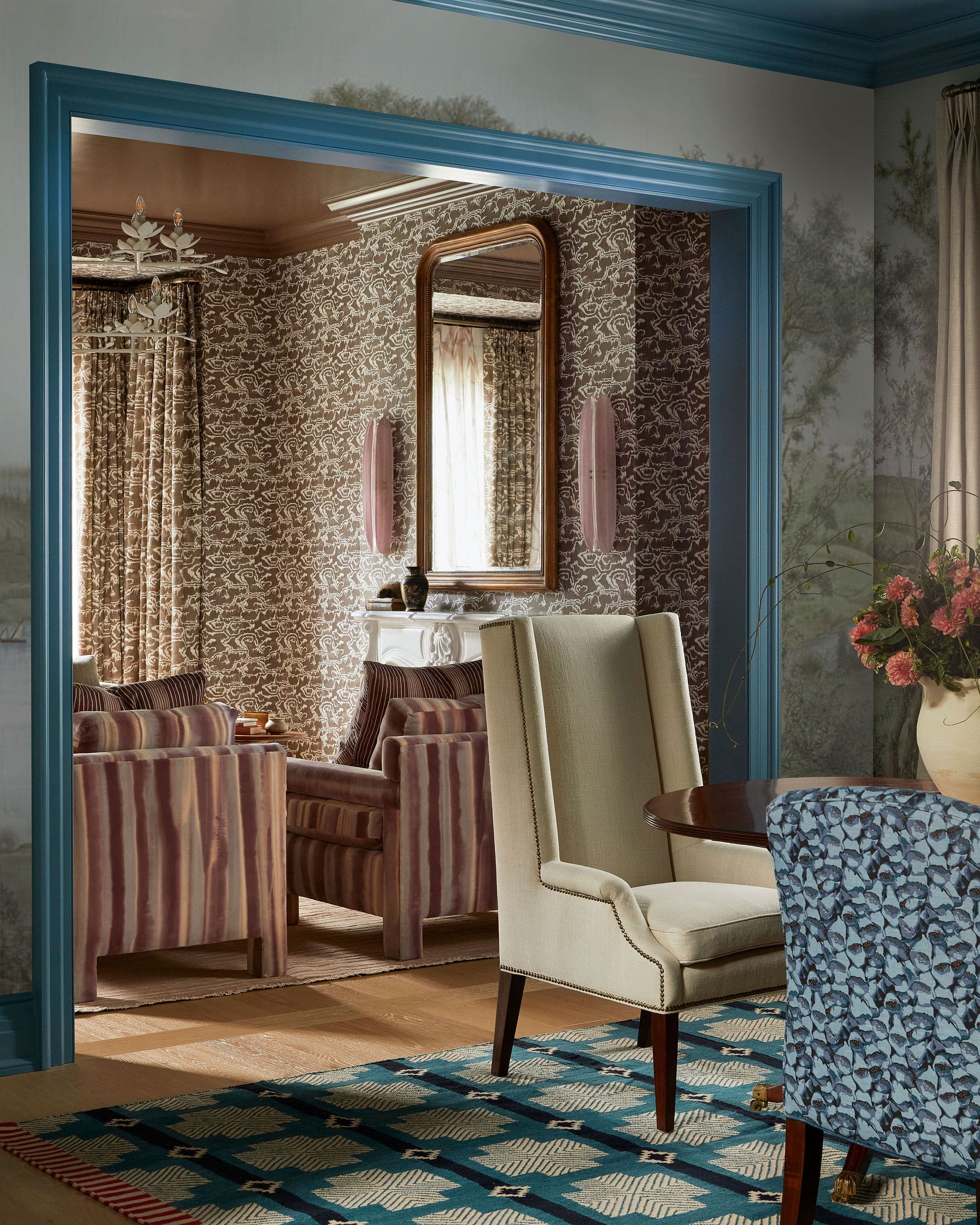 Interior living room seen through a blue-framed doorway, patterned wallpaper, cream upholstered chairs, and a geometric rug.
