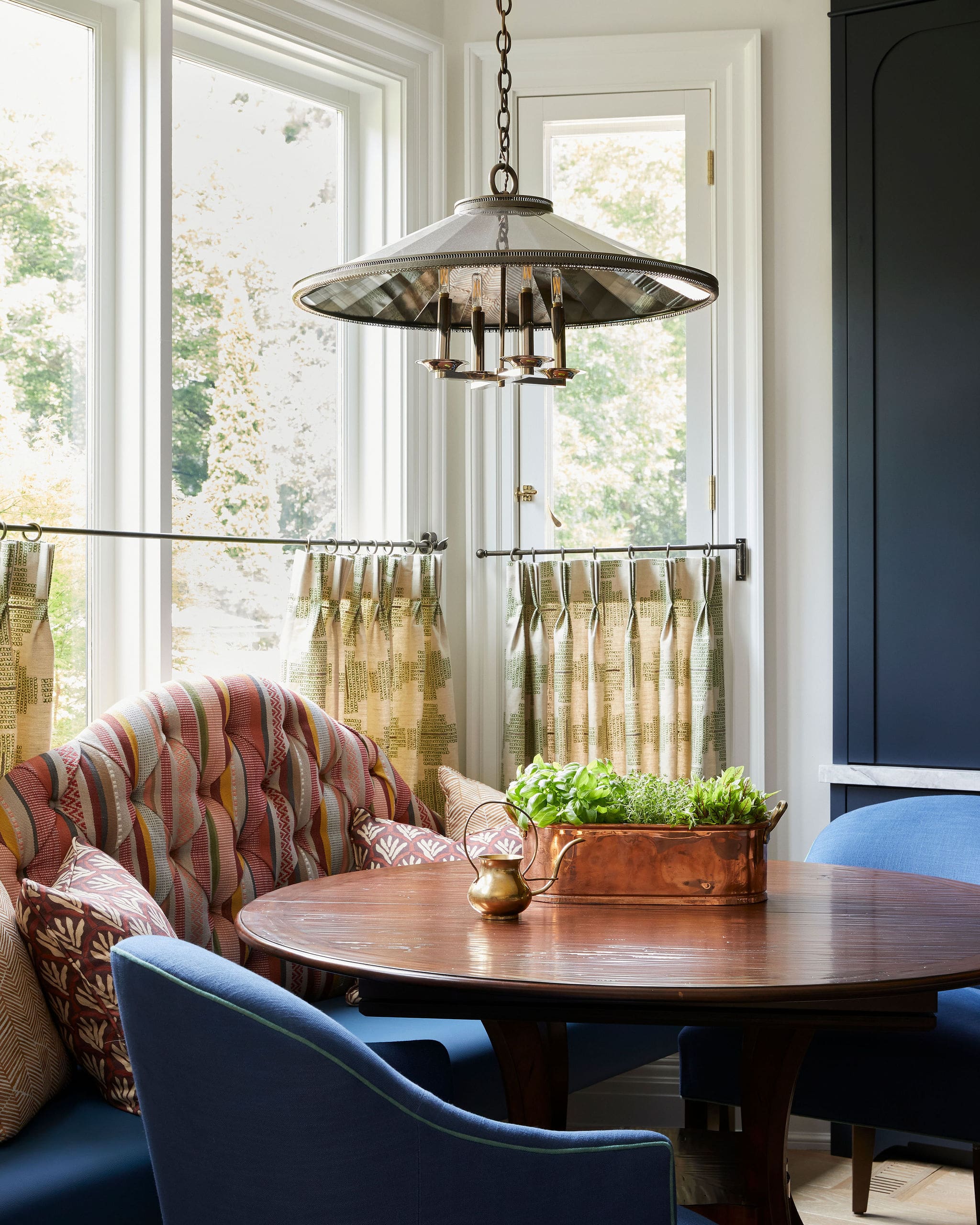 Cozy dining area with a dark blue upholstered chair, round wooden table, and a copper planter with herbs in front of large windows under a hanging light fixture