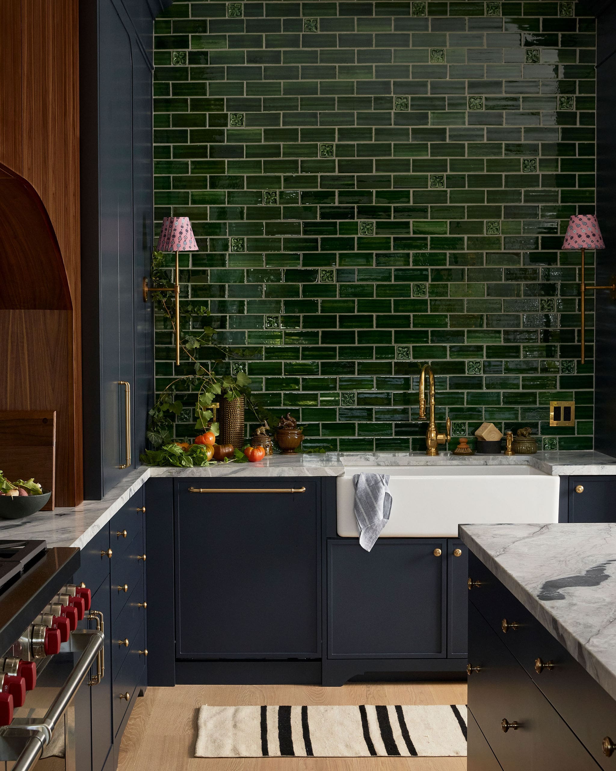 Dark navy kitchen with brass hardware, glossy green brick-style tile backsplash, and a marble countertop; pumpkins and greenery sit along the counter by a white sink area.