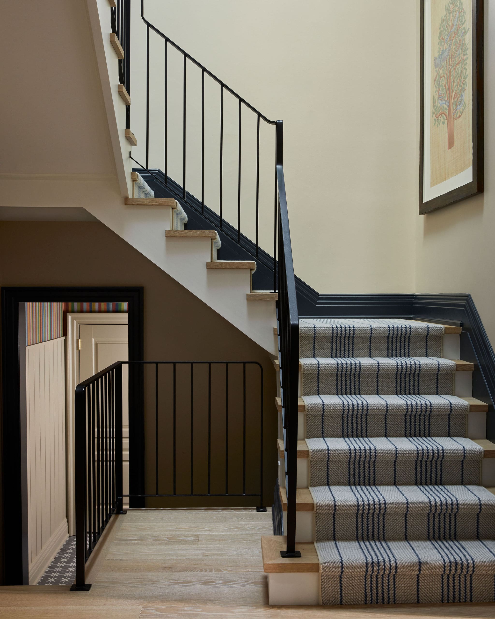Interior staircase with beige walls, black metal railing, and light wood treads covered by a striped carpet runner on a rise to the next floor.