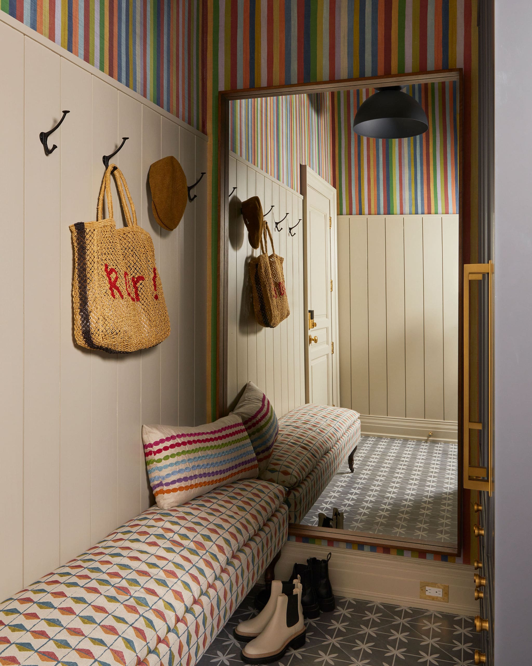 Narrow entryway with cream paneled walls, multicolored striped wallpaper, hooks holding a woven bag and hat, a mirror, and a bench with rainbow-striped cushions and boots on a patterned floor.