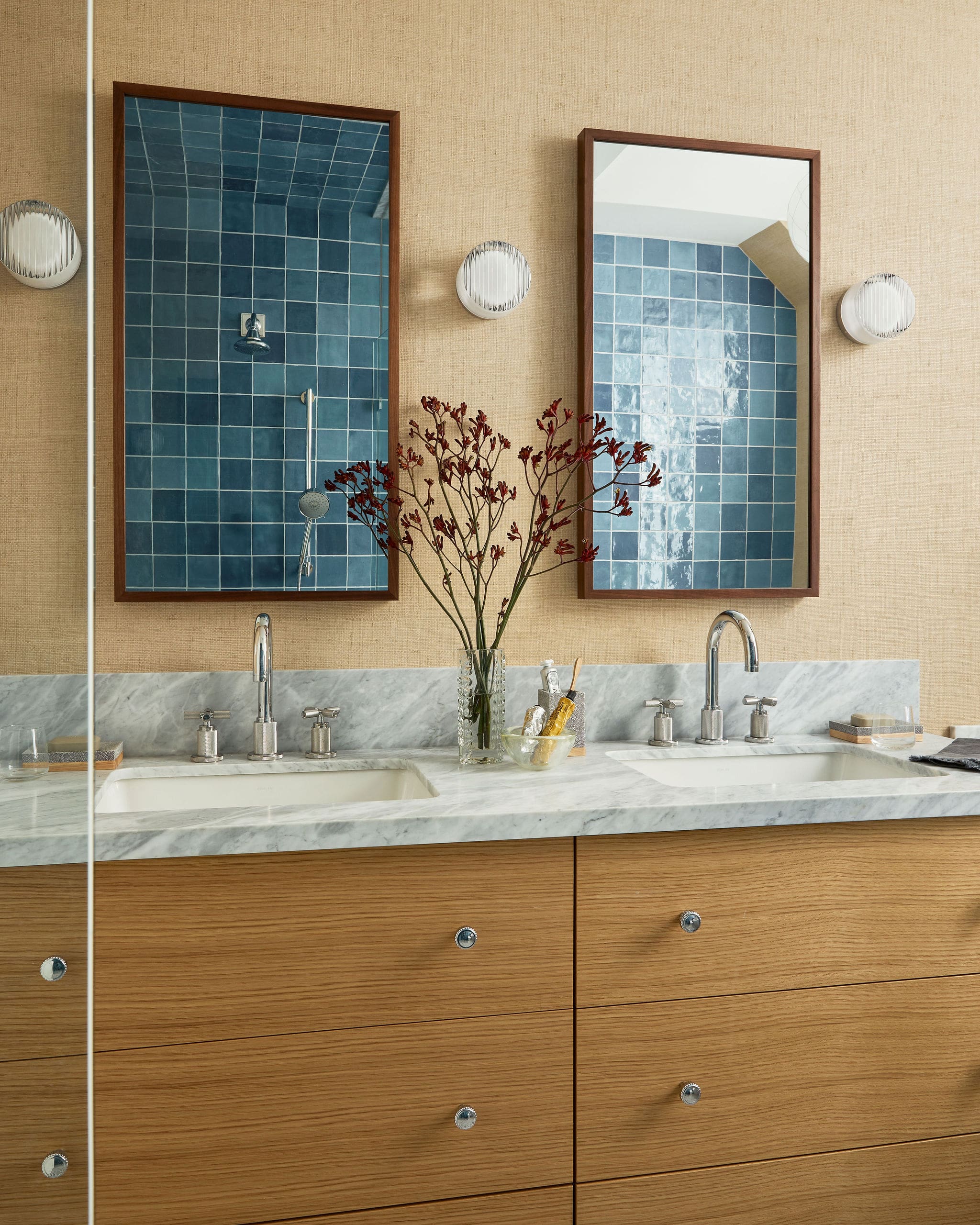 Dual framed mirrors above a marble double sink vanity, blue tile shower reflected in the windows, and a vase with red branches on the counter
