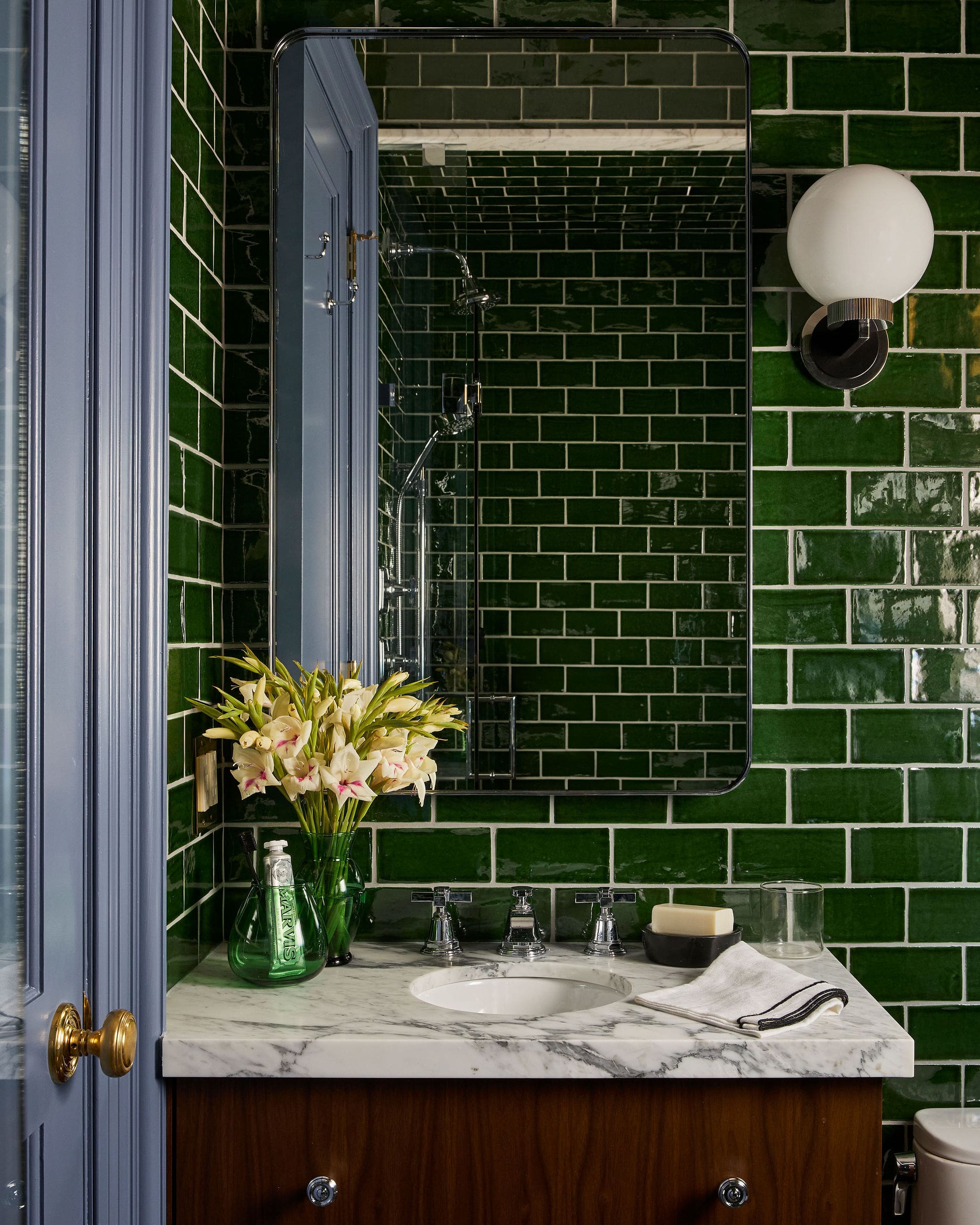 Green-tiled bathroom with a marble countertop vanity, mirror, and pale flowers in a vase.