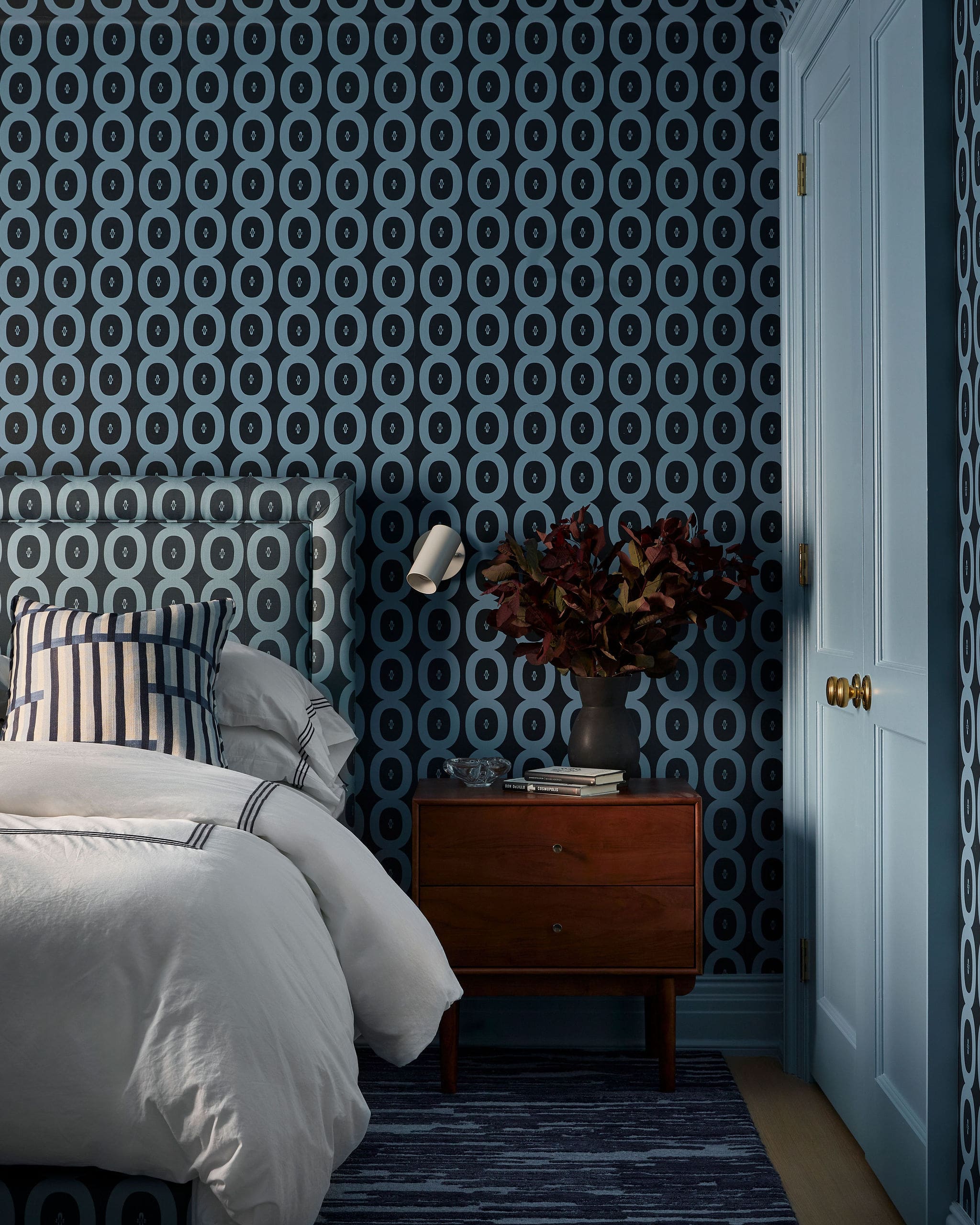 Bedroom corner with blue circular-pattern wallpaper, white bedding with striped pillows, a wooden nightstand, a plant, and a light blue door.