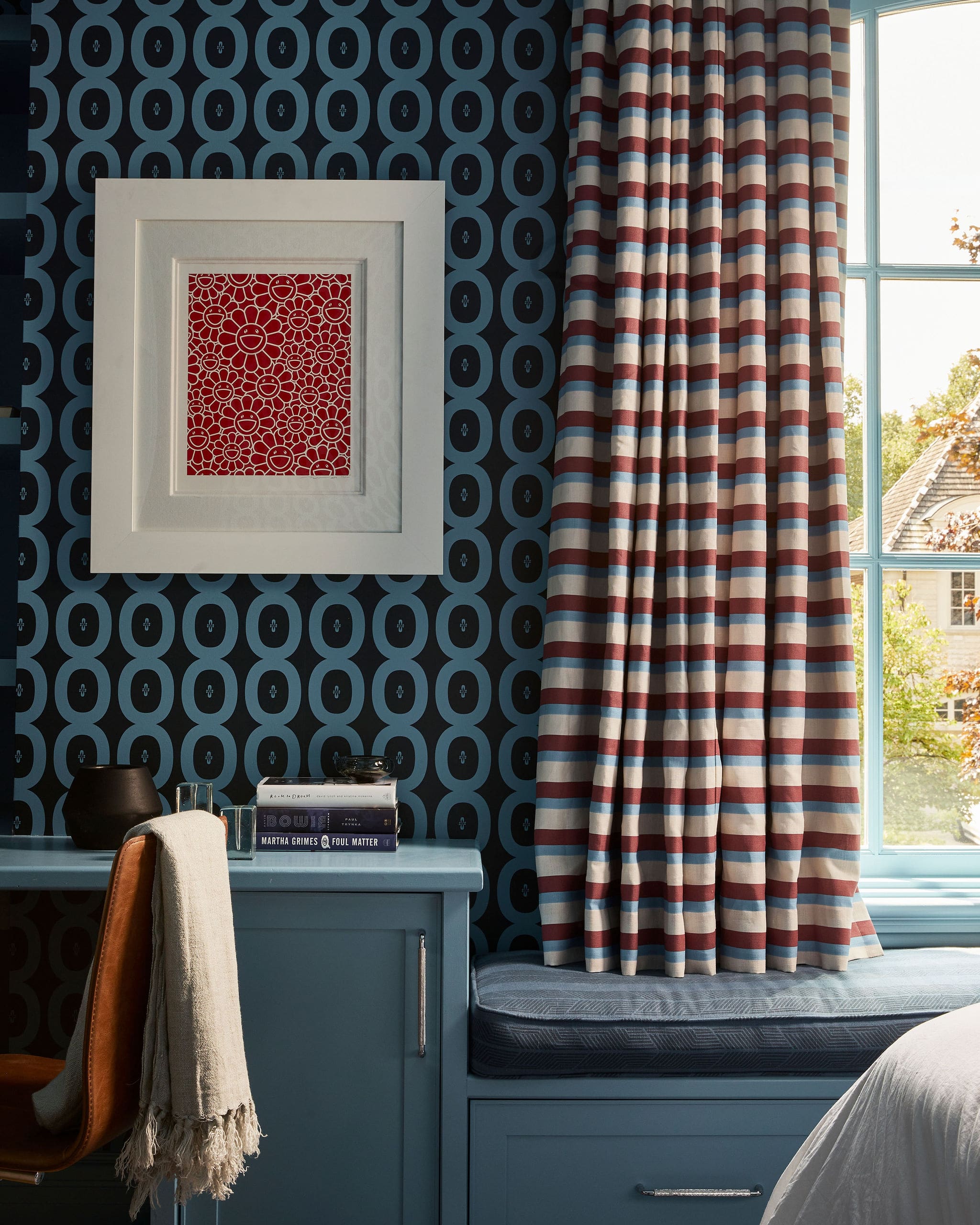 Blue desk or cabinet against dark blue wallpaper with a repeating teal circle pattern, a white frame holding a red floral print, and red, white, and blue striped curtains by a window.