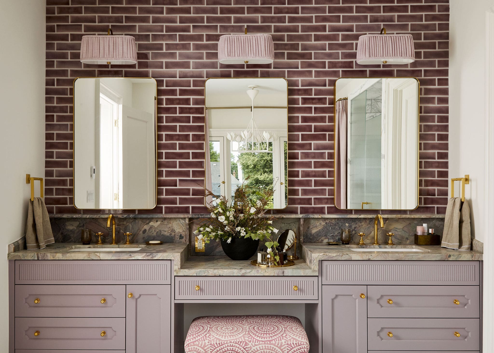 Lavender vanity with marble countertop, three gold-trimmed mirrors, and pink lampshades against a brick wall. Includes flowers and a patterned stool.