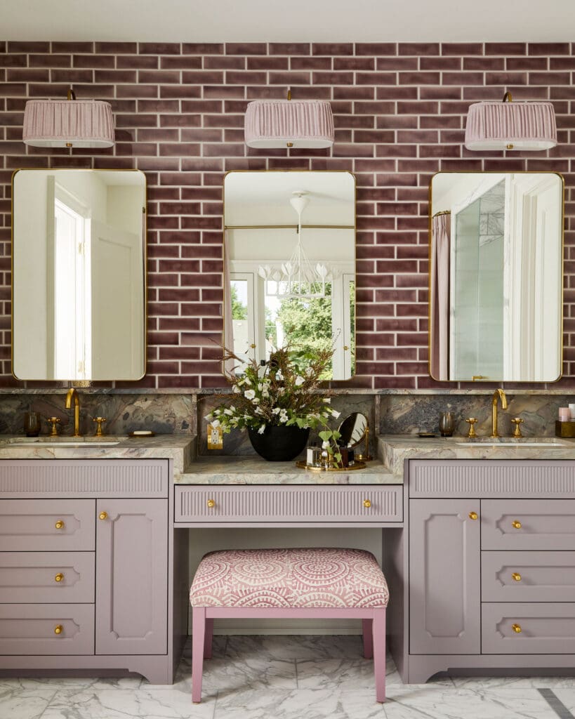 Lavender bathroom vanity with marble countertop, three rounded mirrors, brass hardware, brick backsplash, and pink patterned stool in front.