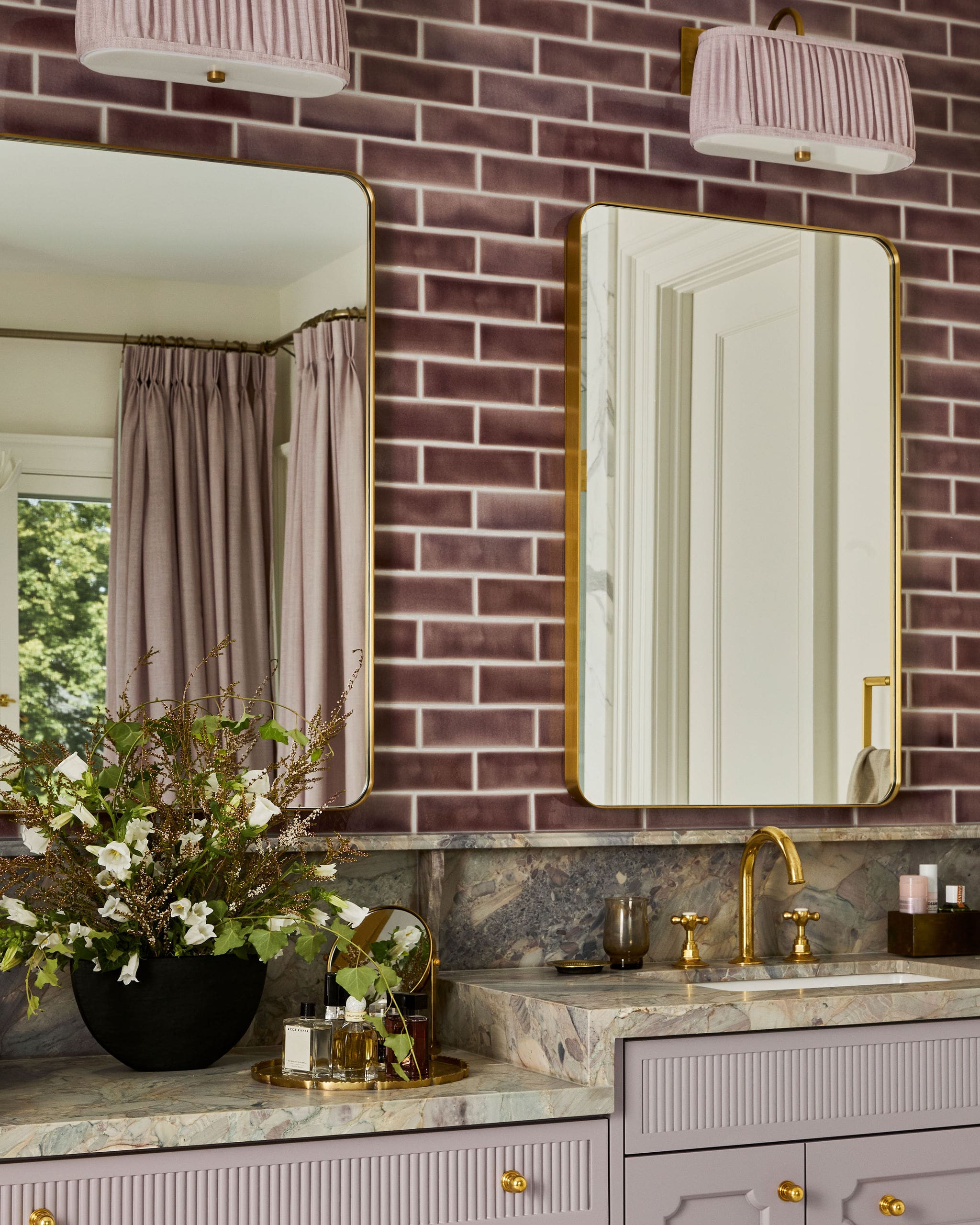 Lavish bathroom vanity with two gold-framed mirrors, lavender cabinetry, marble countertop, pink pleated lamps, and a large floral arrangement.