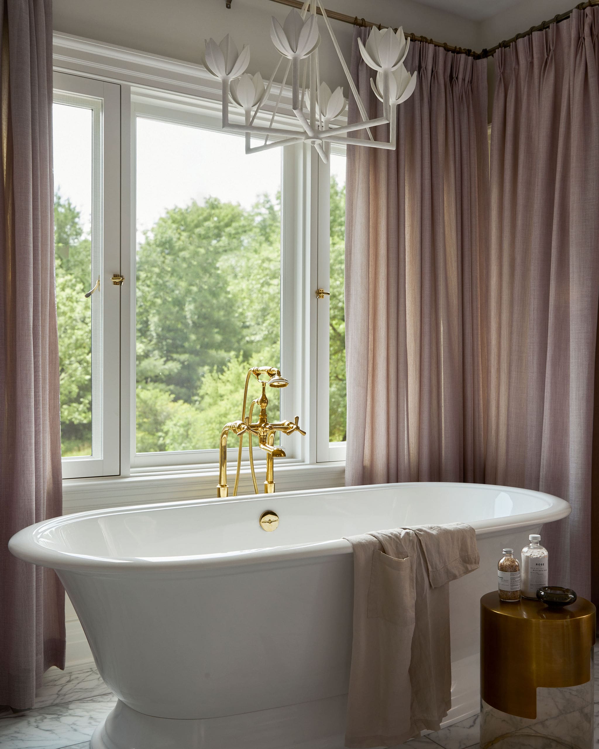 White freestanding bathtub with gold faucet and handheld shower, by a window with pink curtains and marble floor. Outside greenery visible.