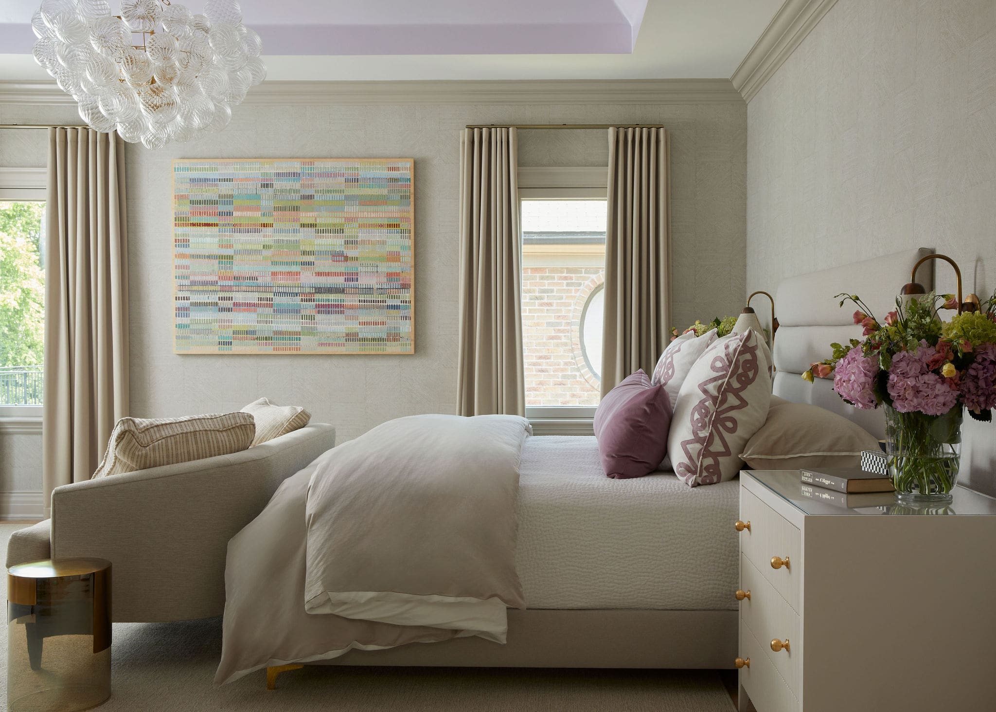 Cozy bedroom featuring a large bed with pale bedding, colorful abstract wall art, and a bouquet on a white dresser.