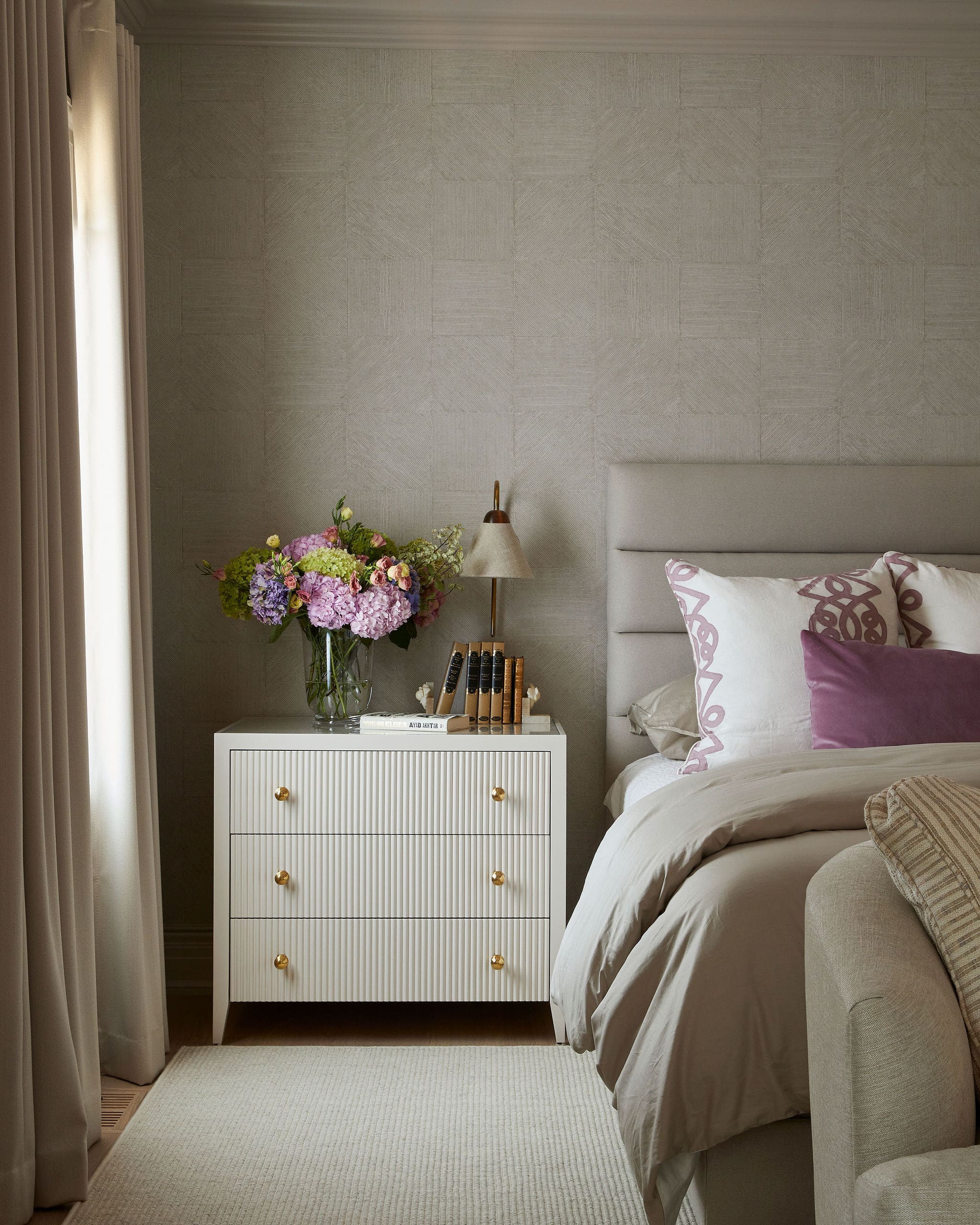 Neutral-toned bedroom with a white ribbed dresser, fresh flowers in a glass vase, a brass-topped lamp, and a bed with beige bedding and pink patterned pillows.