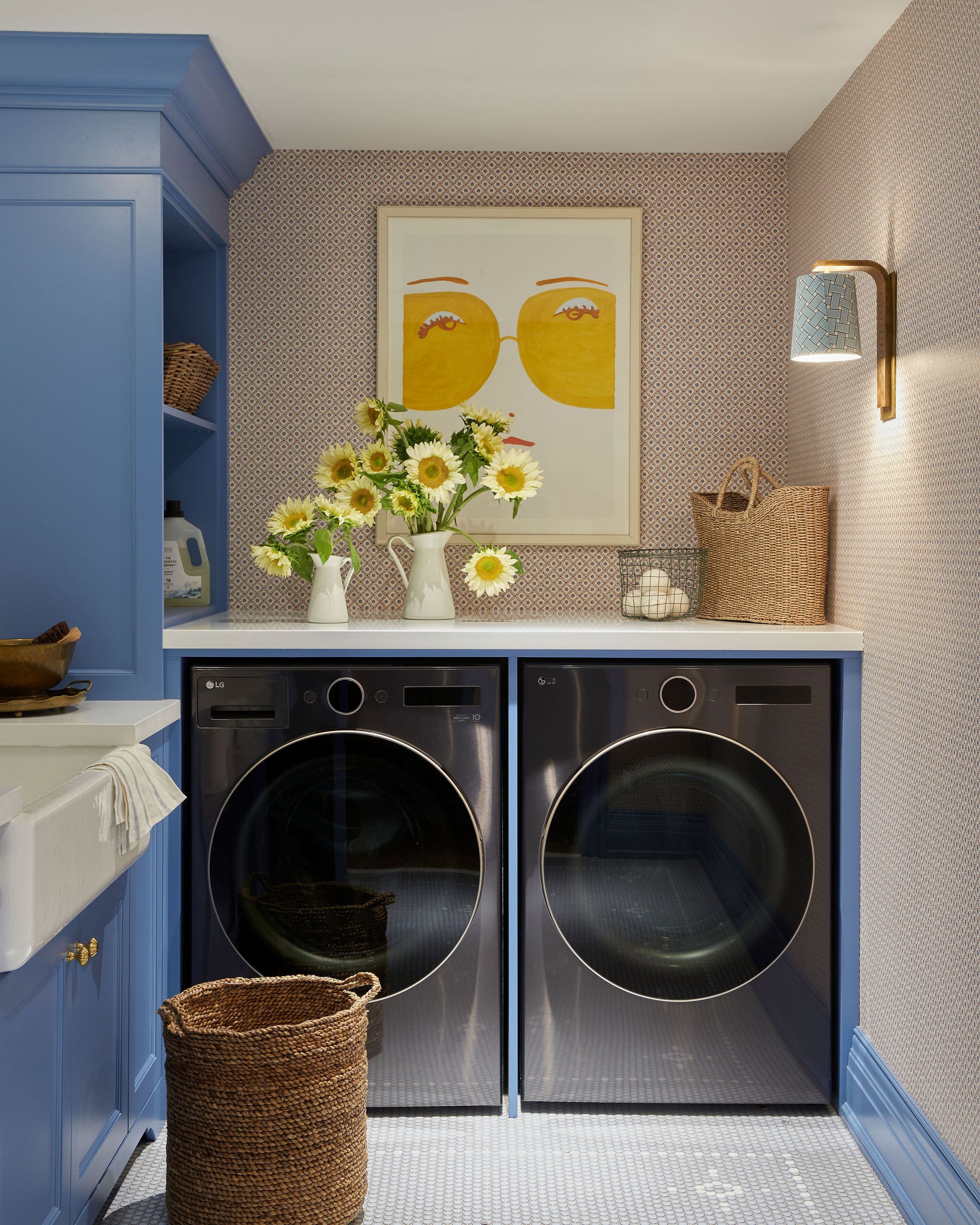 Blue laundry cabinets frame a pair of front-loading washer and dryer, with a sunflower vase and a yellow framed print on the wall behind.