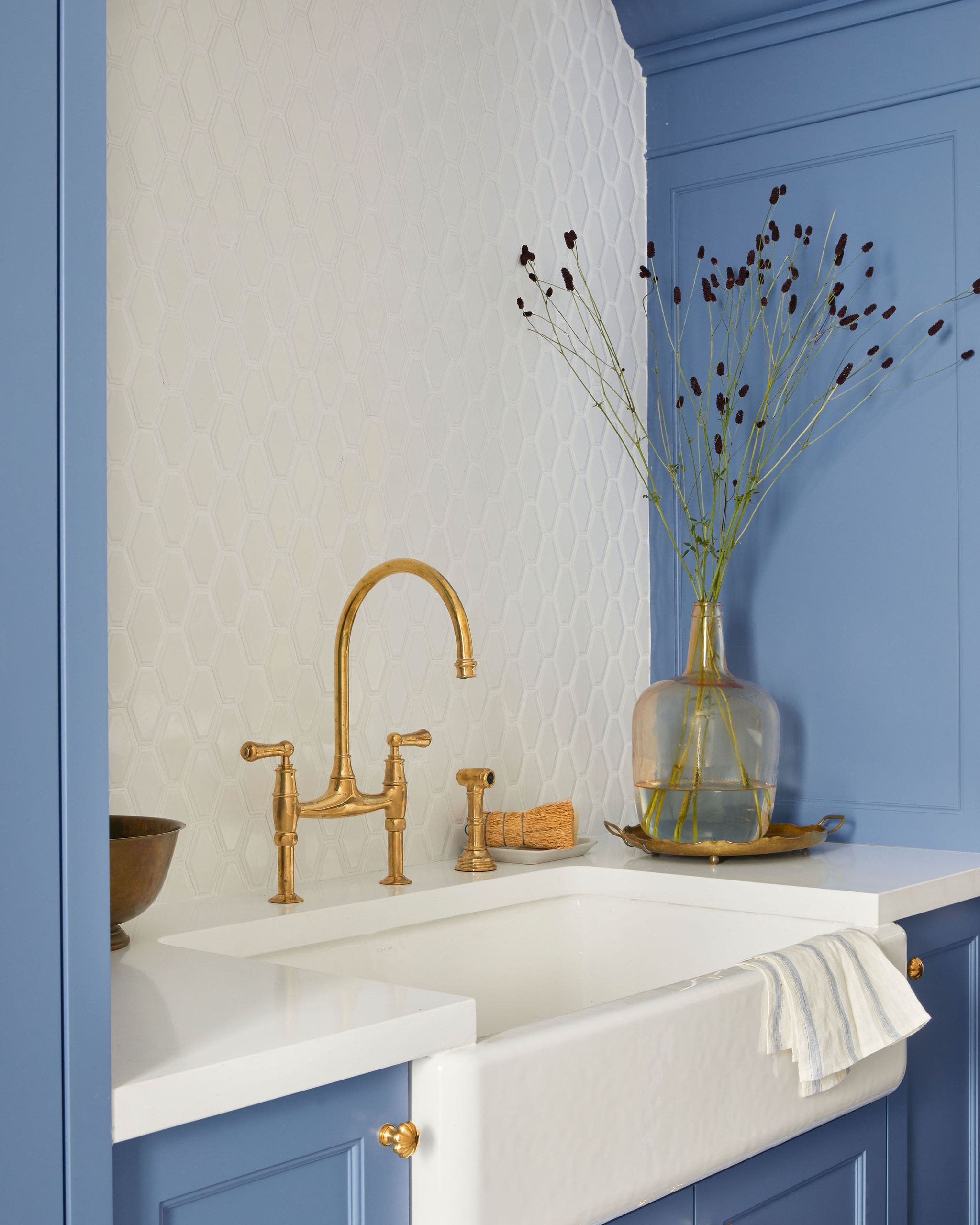 Blue bathroom vanity with a gold faucet, white countertop, and a vase of tall dried flowers against a blue paneled wall with hex tile backsplash.