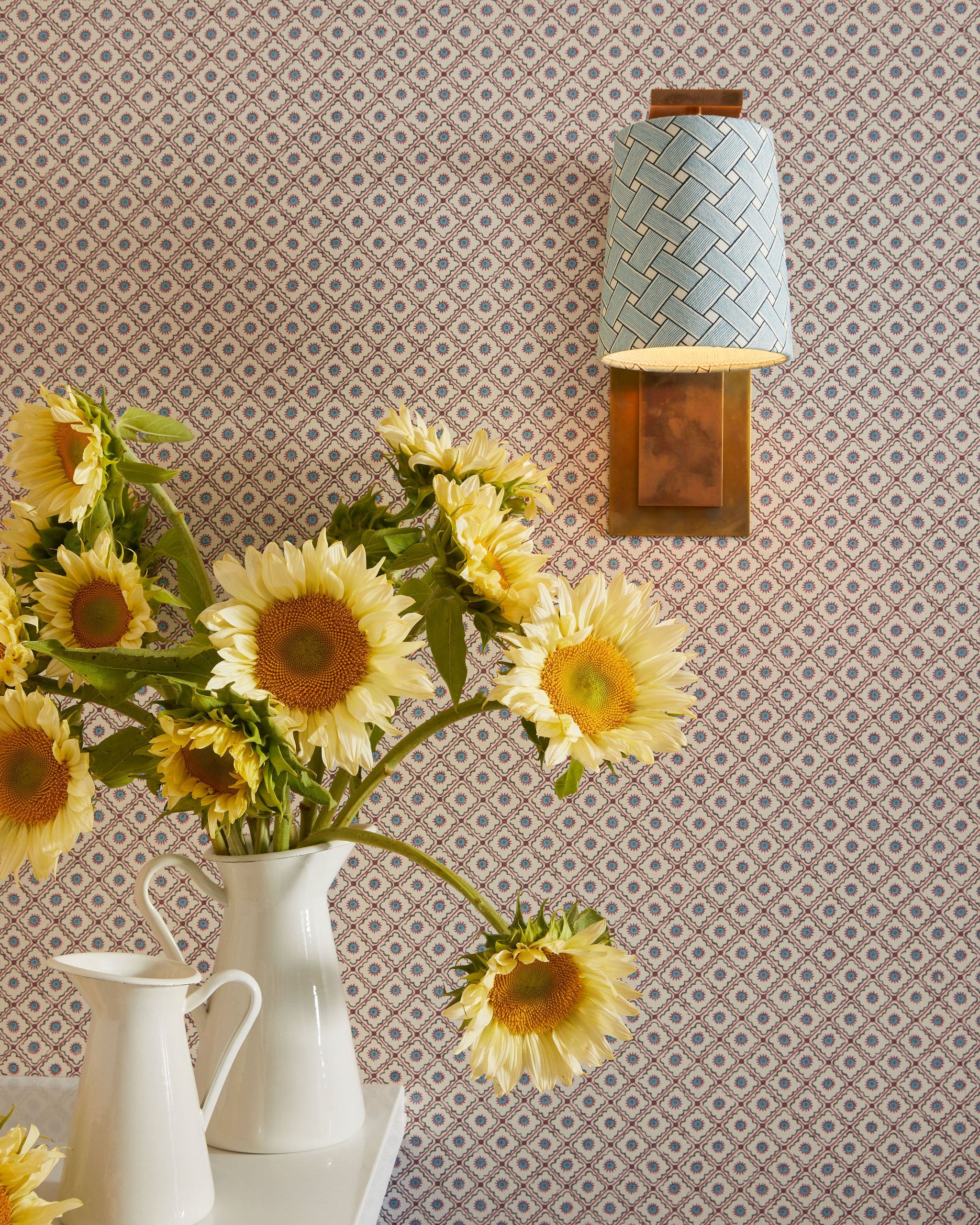 Sunflowers in white ceramic pitchers arranged on a white table with a patterned wallpaper backdrop (geometric diamonds).