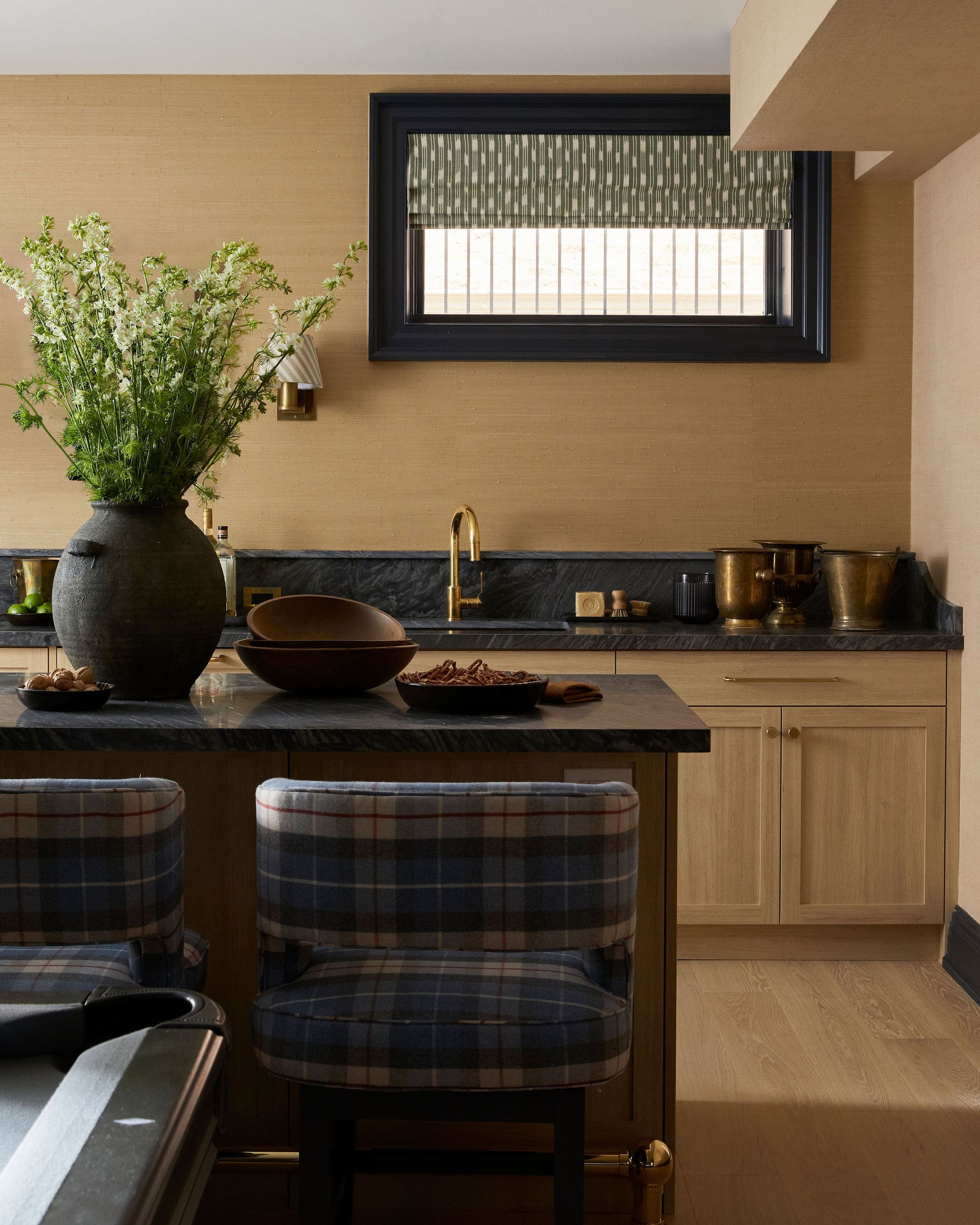 Large kitchen island with black marble countertop, gold faucet, and plaid-upholstered stools facing away from the camera; a vase of white flowers sits on the island.