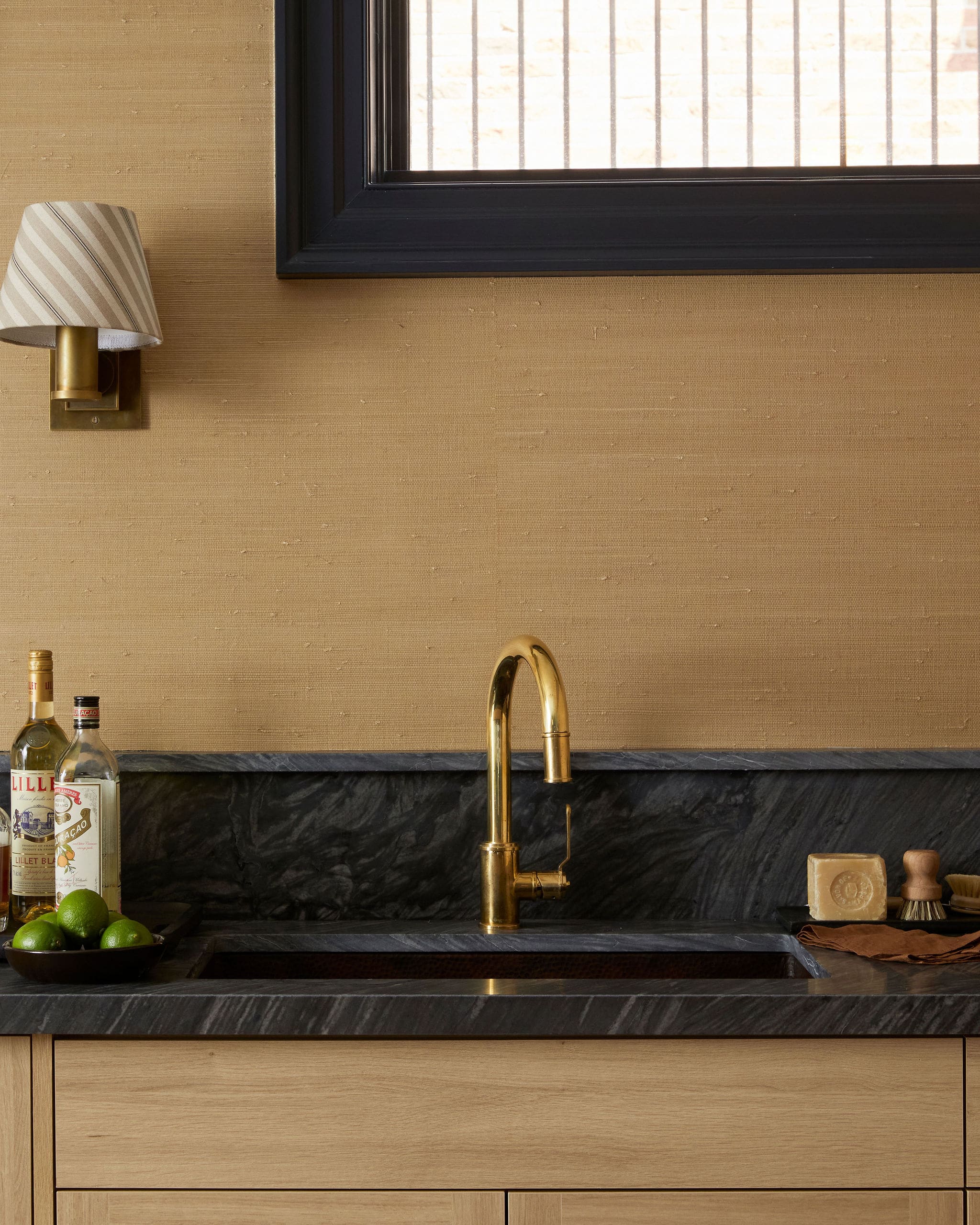 Gold faucet over a dark marble counter with a built-in sink, beige textured wall, and a striped wall lamp on the left.