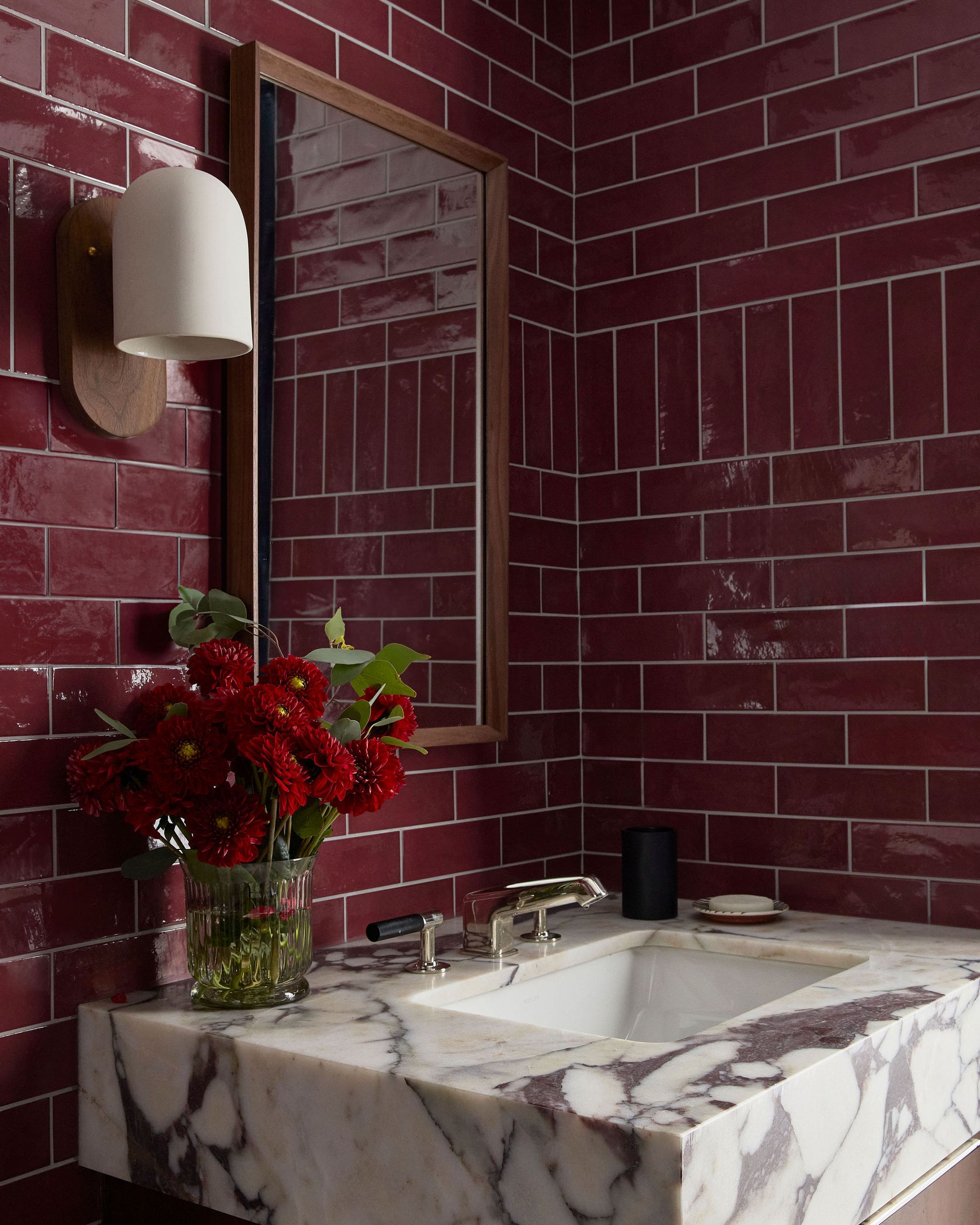 Marble bathroom sink with a glass vase of red flowers, against a burgundy tile wall and a wooden-framed mirror.