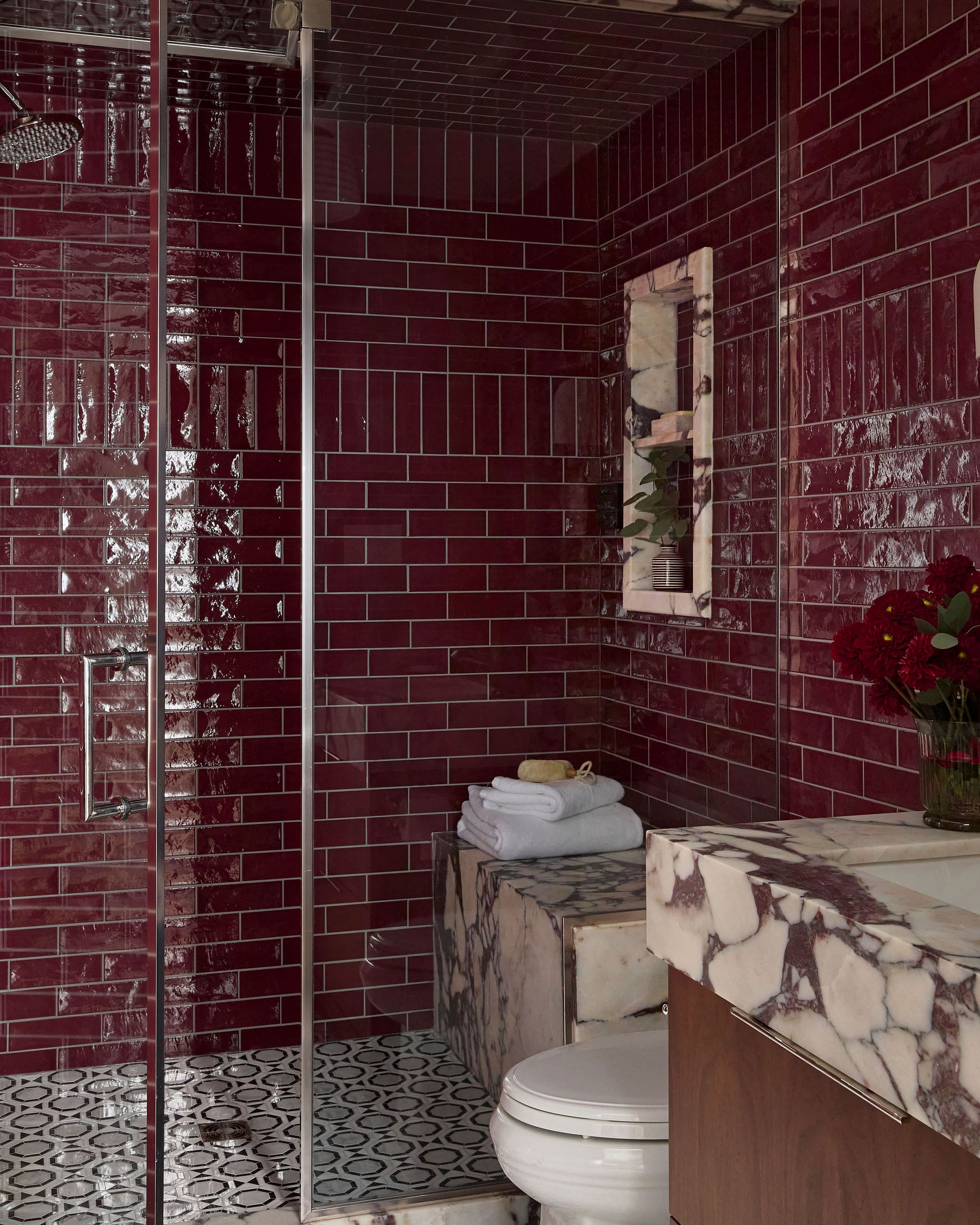 Luxurious bathroom with glossy burgundy subway tiles, glass shower enclosure, and marble vanity top with towels and red flowers nearby