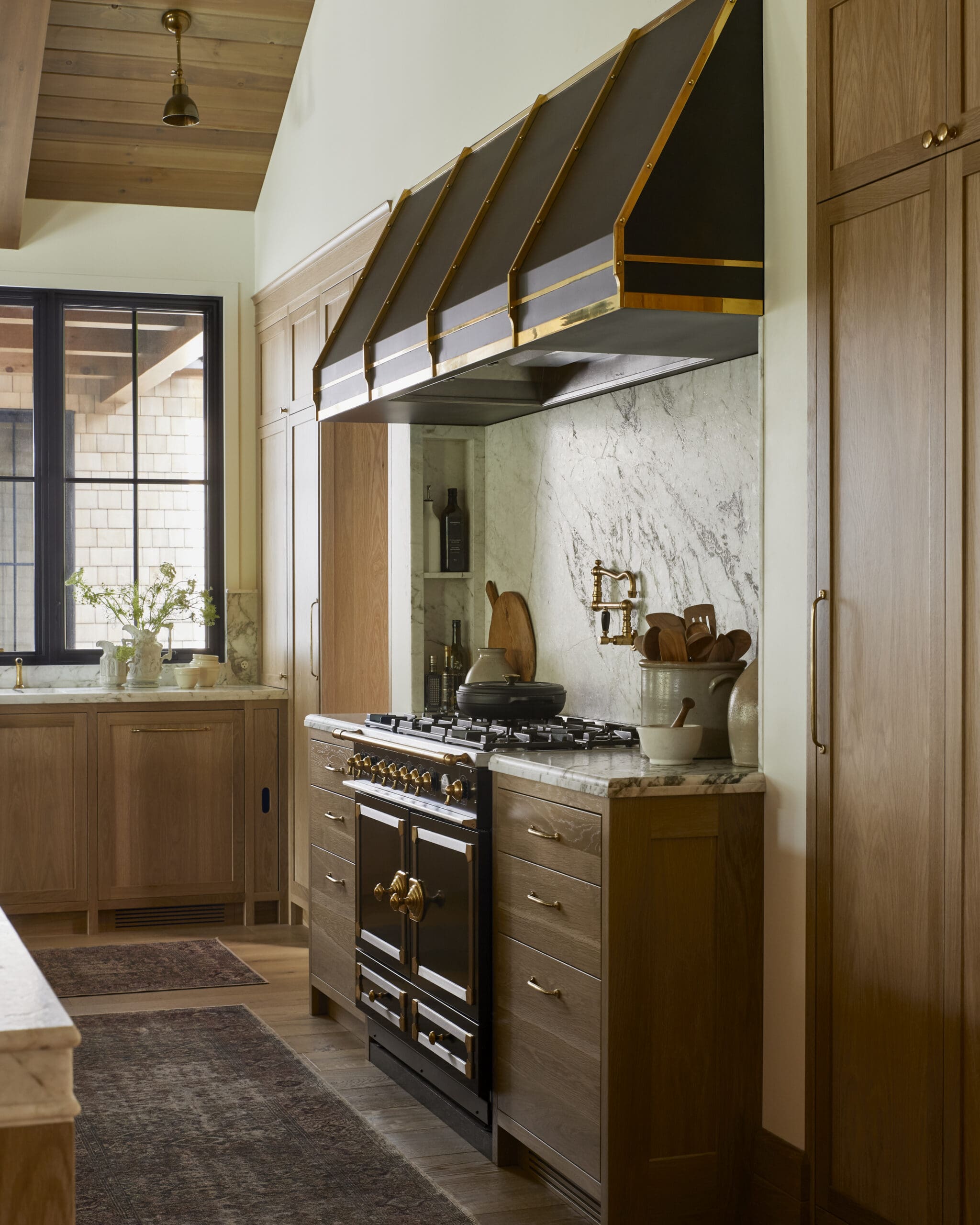 Kitchen with a black range and brass-trimmed hood against marble backsplash and wooden cabinets.