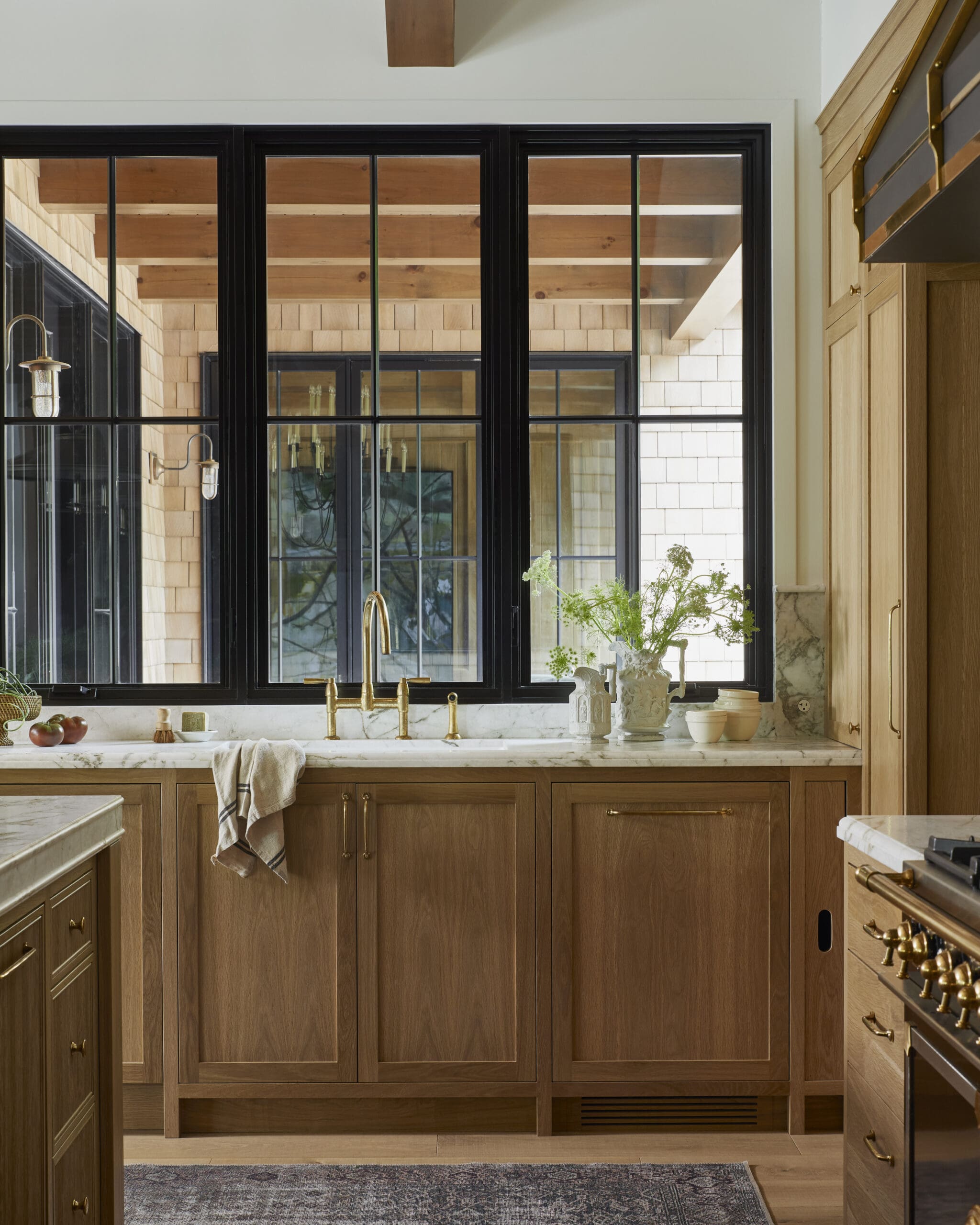Wooden kitchen cabinets and drawers beneath a marble countertop with a large black-framed window wall and brass faucet fixtures.