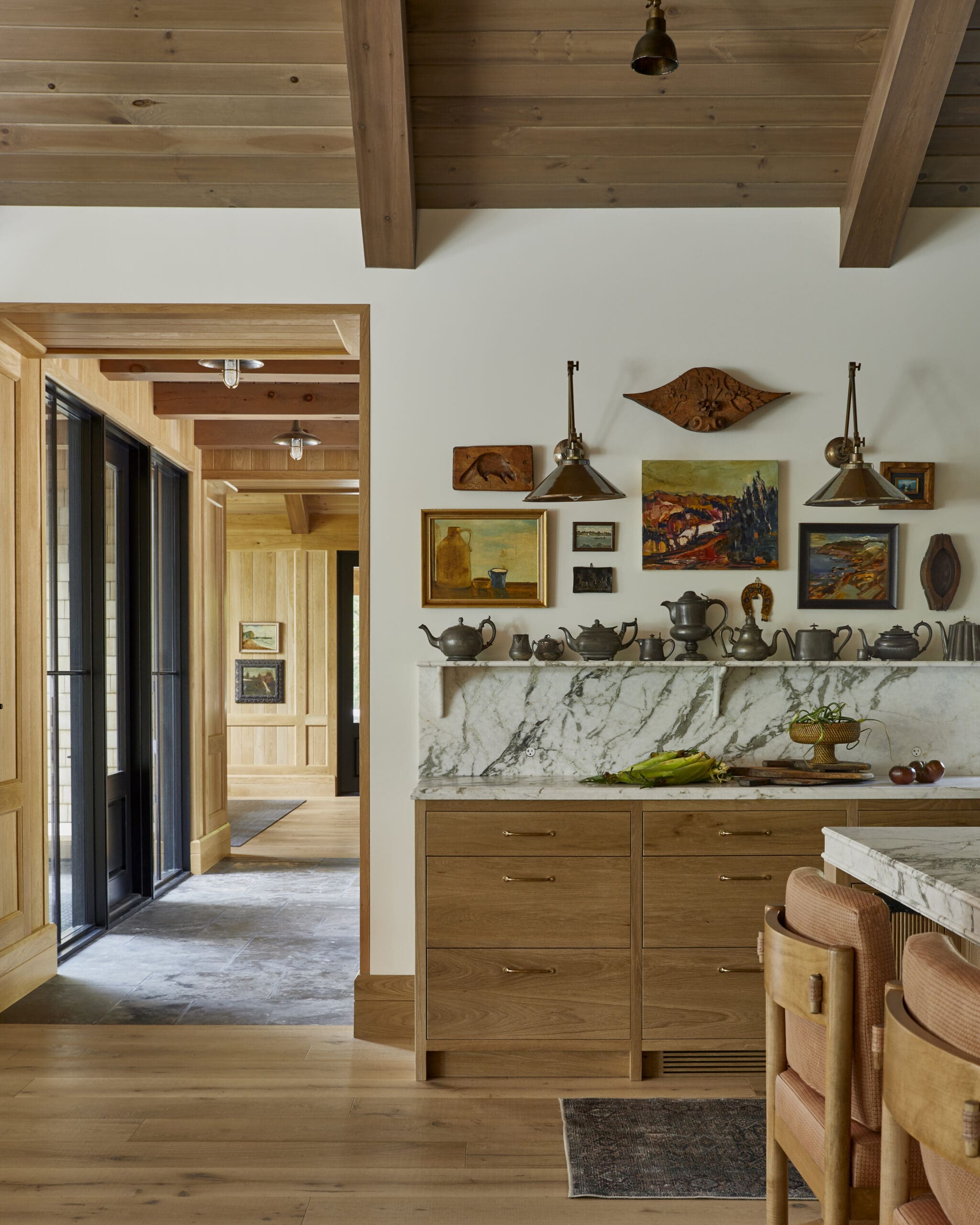 Rustic kitchen with marble island, wooden cabinets, and a wall gallery of paintings under brass pendant lights.