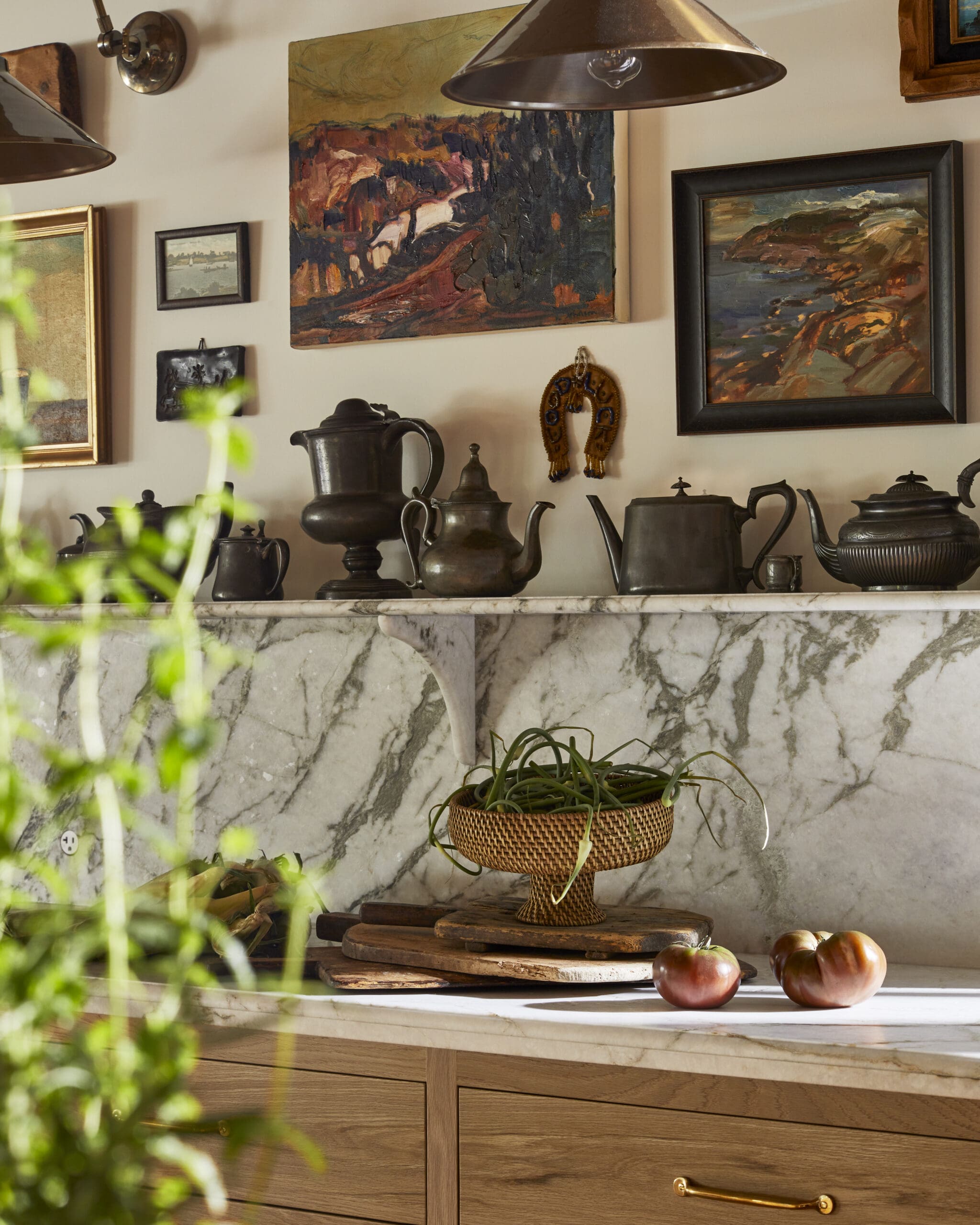 Shelf displaying antique teapots and framed artwork over a marble surface, with a woven basket of plants and apples nearby