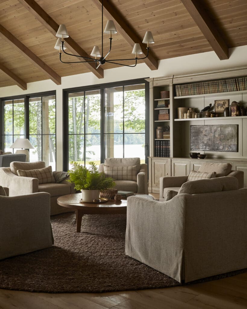 Living room with a wooden vaulted ceiling and black chandelier, overlooking large glass doors and built-in shelves filled with books and decor.