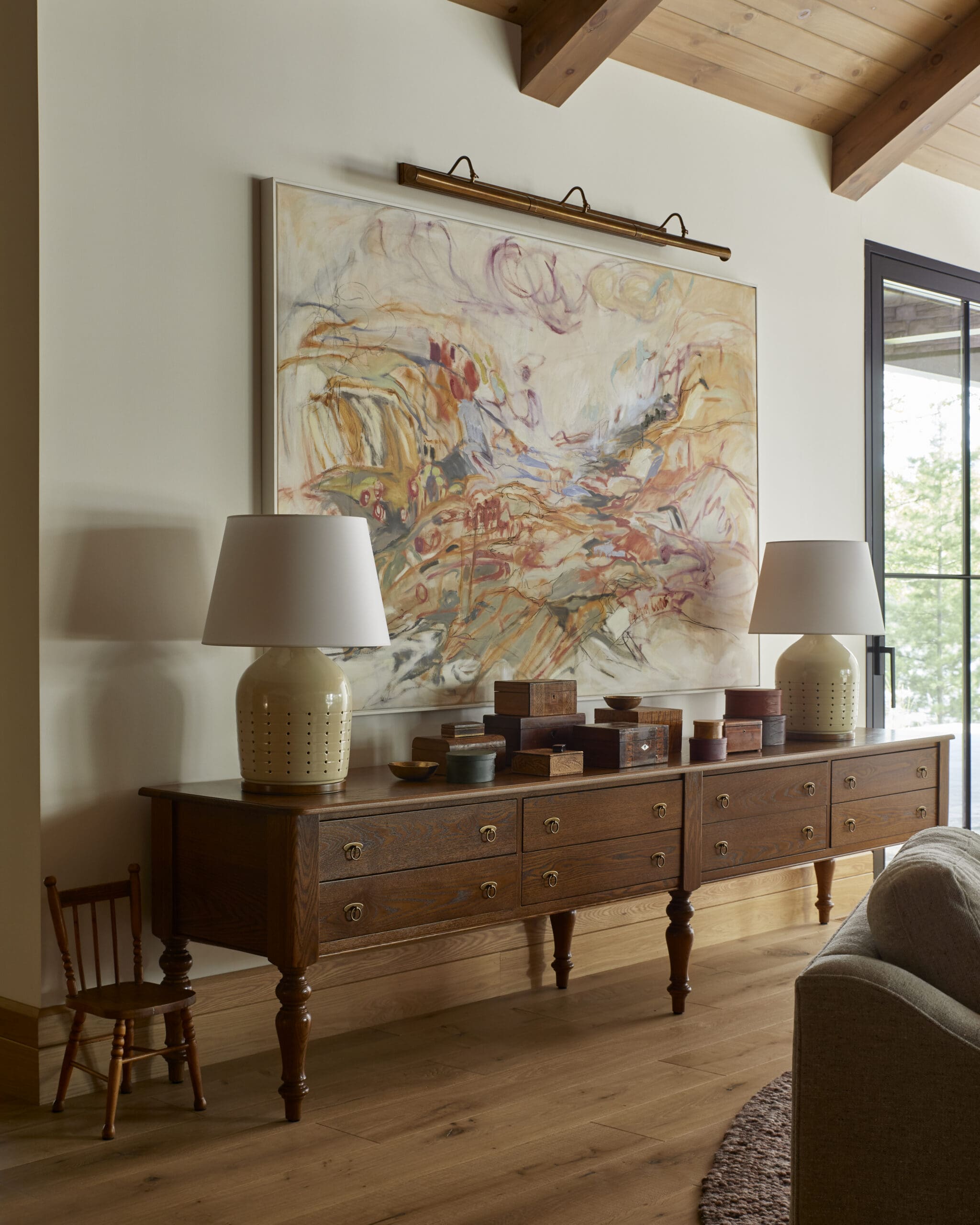 Abstract artwork hung above a long wooden console with brass drawer pulls, flanked by two beige lamps.